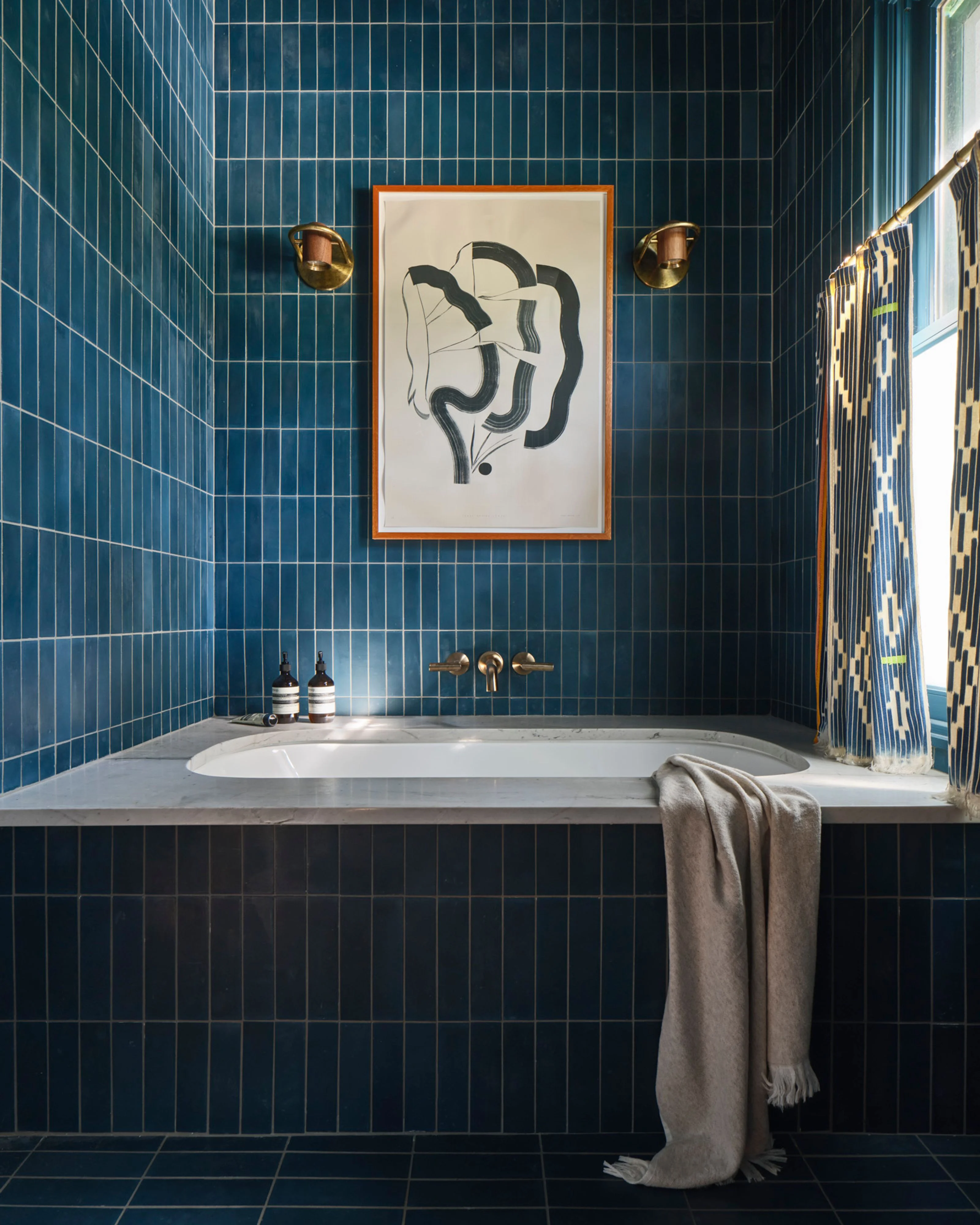 A modern bathroom features floor-to-ceiling deep teal vertical subway tiles with white grout lines. A white oval soaking tub sits centered beneath an abstract line-drawing artwork in an orange frame, flanked by brass sconces and wall-mounted faucet fixtures, with a beige linen towel draped over the edge.