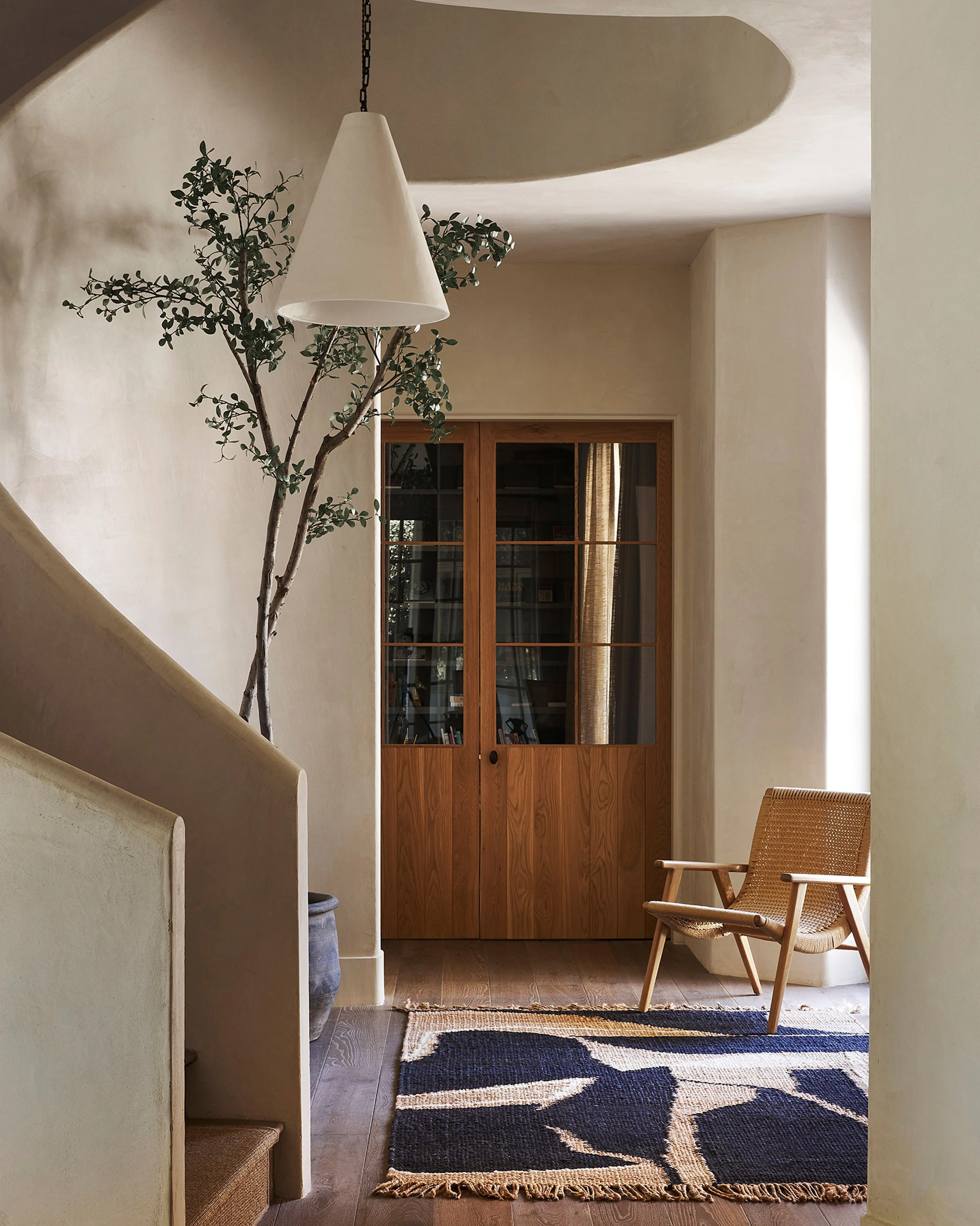 A serene modern interior features curved cream-colored plaster walls with soft natural lighting from a large circular skylight, a minimalist white cone pendant lamp, and warm wood flooring accented by a geometric navy and cream patterned rug. The space includes a wooden-framed glass door, a woven mid-century modern chair, and delicate botanical branches in a vase.