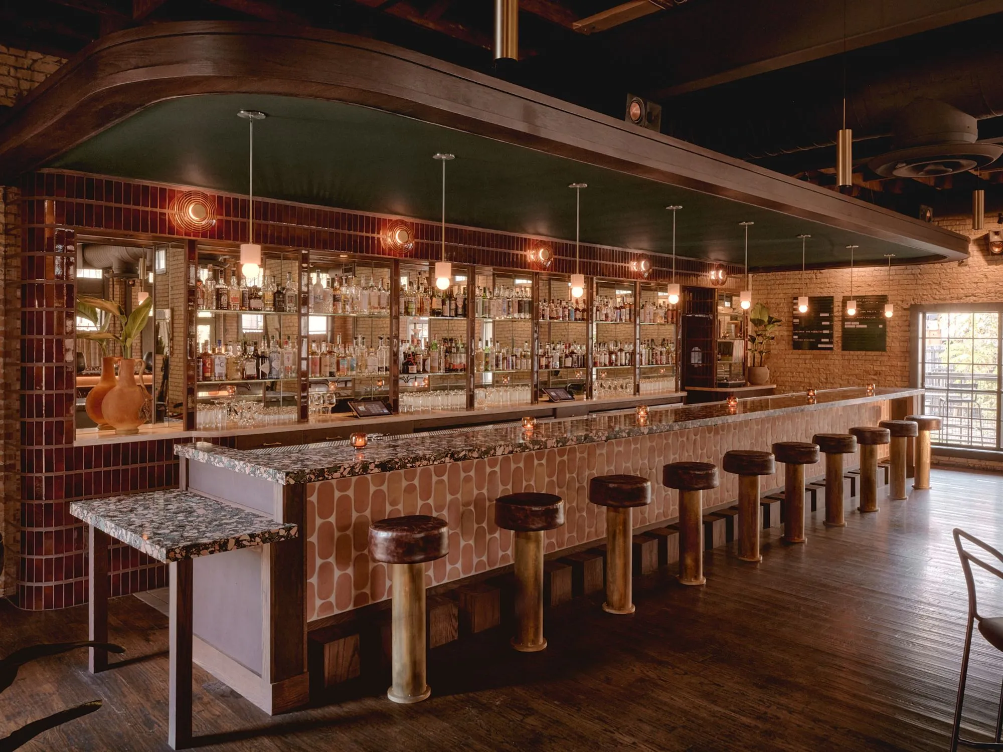 A sophisticated high vibe bar interior featuring warm terracotta brick walls, a curved wooden ceiling soffit, and an extensive backlit liquor display with decorative red tilework. The space showcases a curved bar counter with geometric patterned upholstery, brass-cylindrical bar stools, and pendant lighting that creates an inviting, warm ambiance.