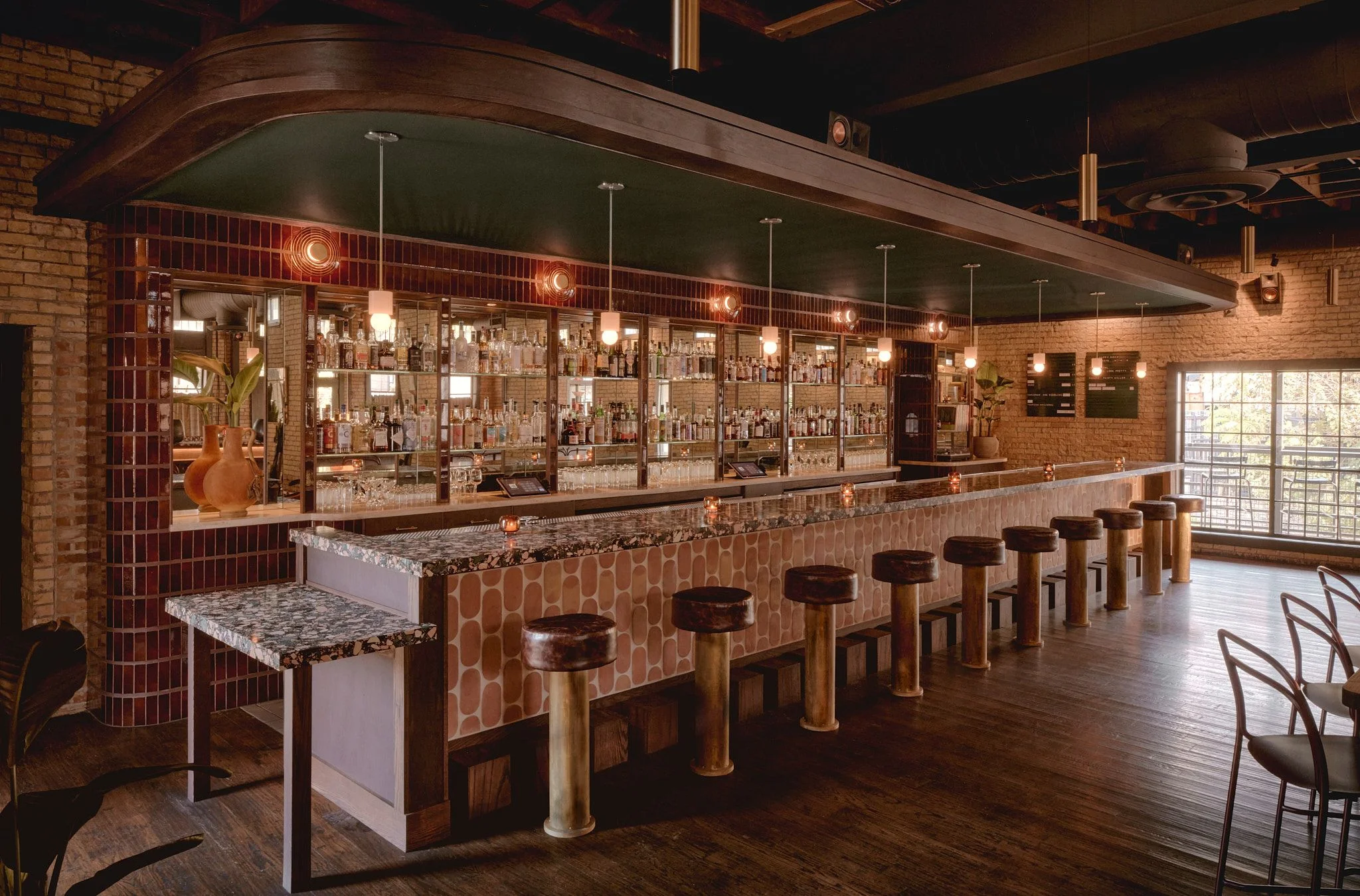 A sophisticated high vibe bar interior featuring warm terracotta brick walls, a curved wooden ceiling soffit, and an extensive backlit liquor display with decorative red tilework. The space showcases a curved bar counter with geometric patterned upholstery, brass-cylindrical bar stools, and pendant lighting that creates an inviting, warm ambiance.