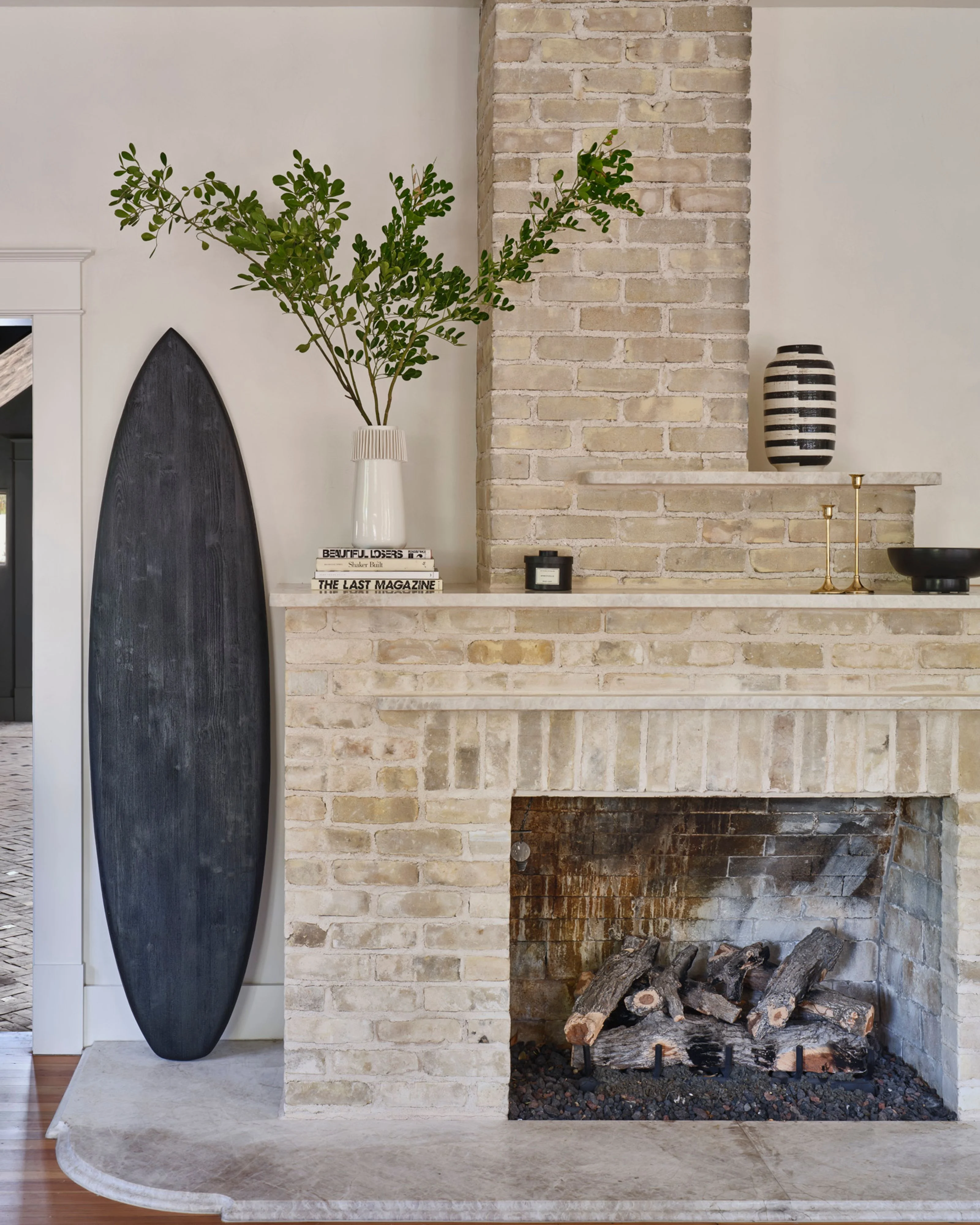 A modern fireplace with a cream-colored brick chimney breast features a working fire with logs, flanked by a minimalist dark wooden oval sculpture and a styled mantel displaying fresh greenery, striped ceramic vessels, stacked magazines, and brass accents.