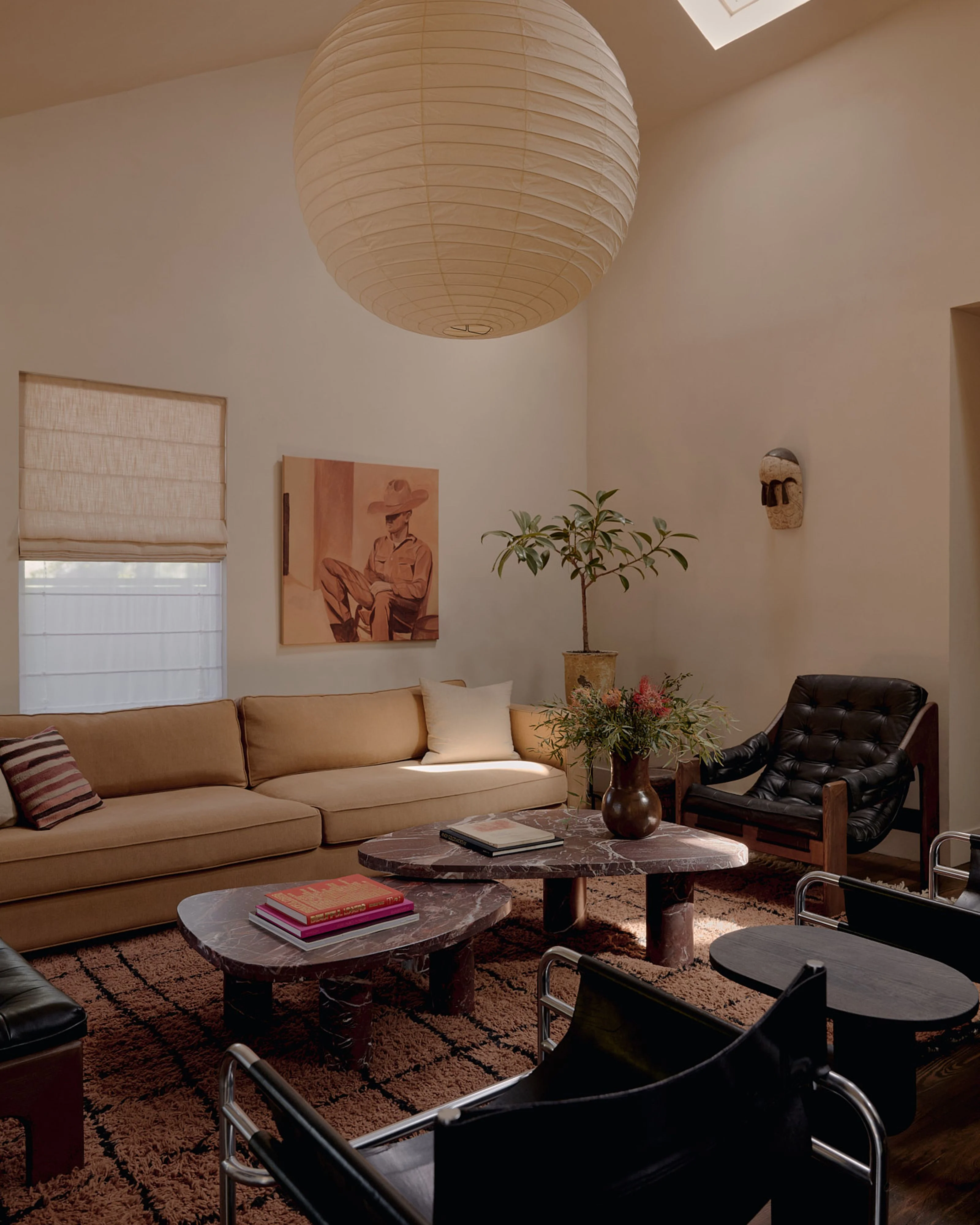 A minimalist living room with warm neutral tones features a large cream paper pendant light suspended from a vaulted ceiling with a skylight, a tan leather sofa, dark marble-topped tables, black leather chairs, and a warm terracotta area rug accented with potted plants and vintage artwork.