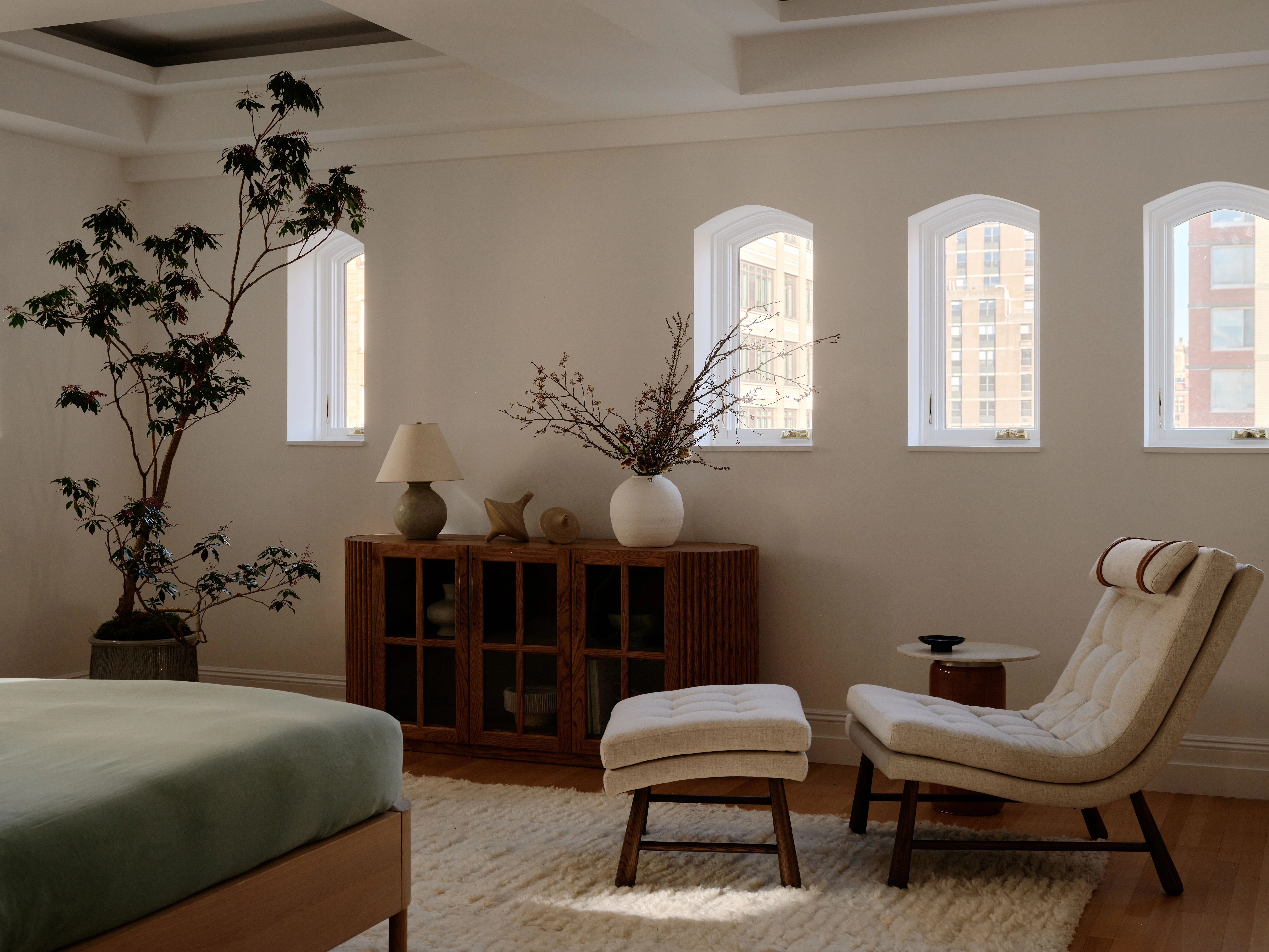 A serene, minimalist bedroom features warm beige walls with three arched white-trimmed windows overlooking urban buildings, mid-century modern furniture including a cream-upholstered lounge chair and matching ottoman, a wooden sideboard displaying botanical elements and a table lamp, and abundant natural light creating a calm, contemporary aesthetic.