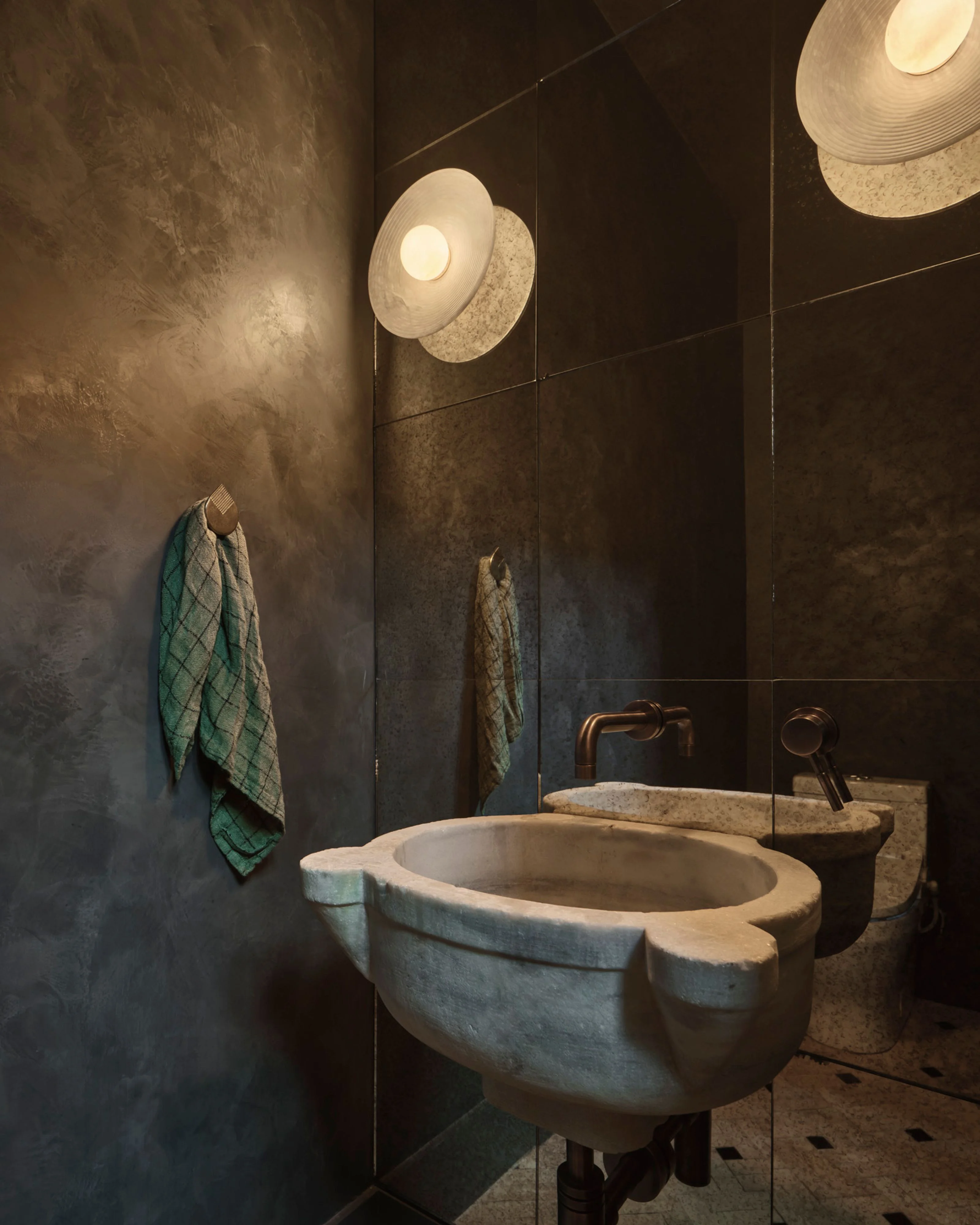 A moody, contemporary bathroom features dark textured walls with a sculptural stone sink, warm pendant lights with woven shades, bronze fixtures, and a green towel, creating an earthy, minimalist aesthetic with dramatic lighting.