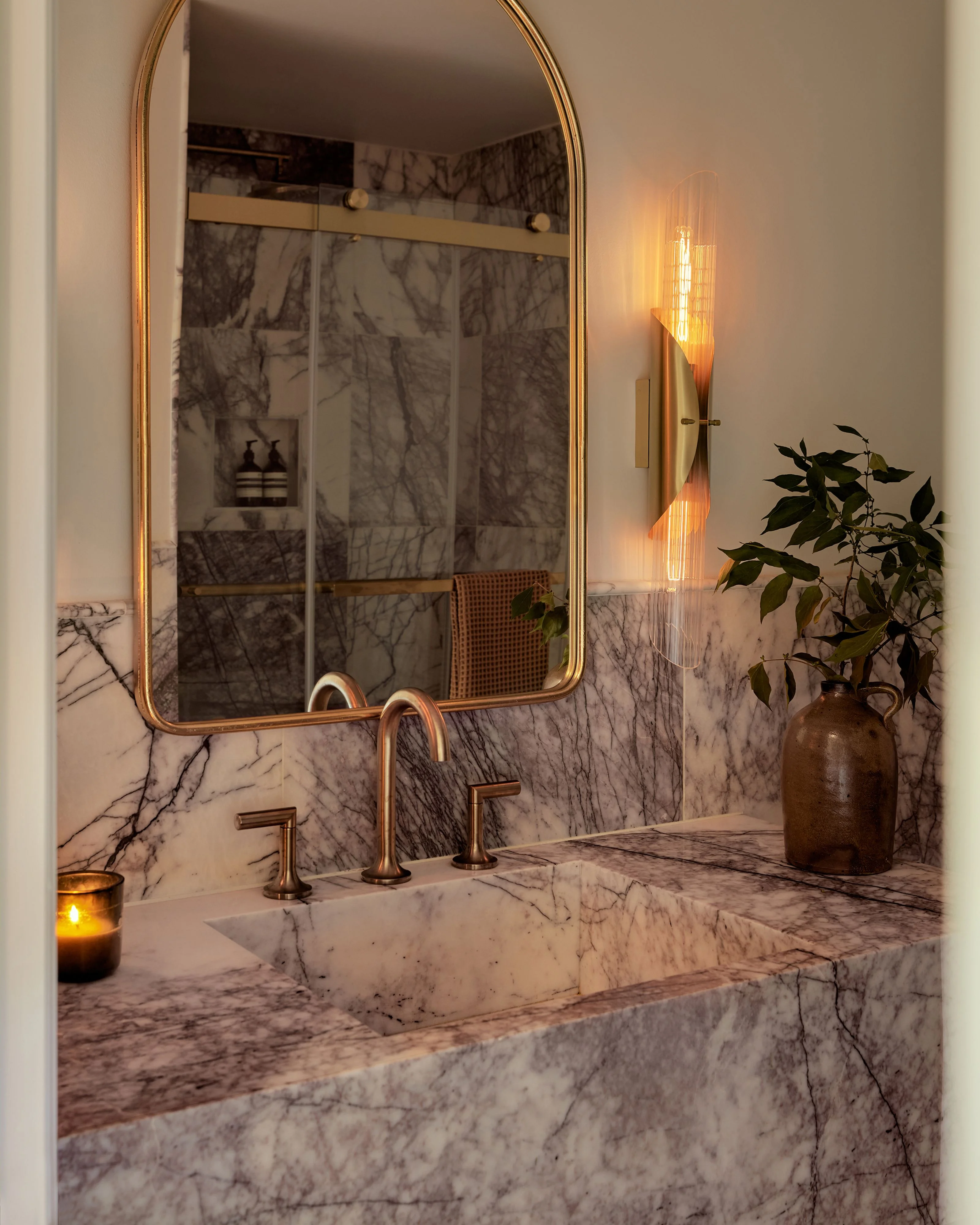 A luxurious modern bathroom features a marble sink counter with brass fixtures and an arched gold-framed mirror, illuminated by a glowing vertical light fixture and warm candlelight, with marble tile walls and a glass shower enclosure visible in the background. A bronze vase with fresh greenery and decorative dried branches complete the serene, spa-like aesthetic.