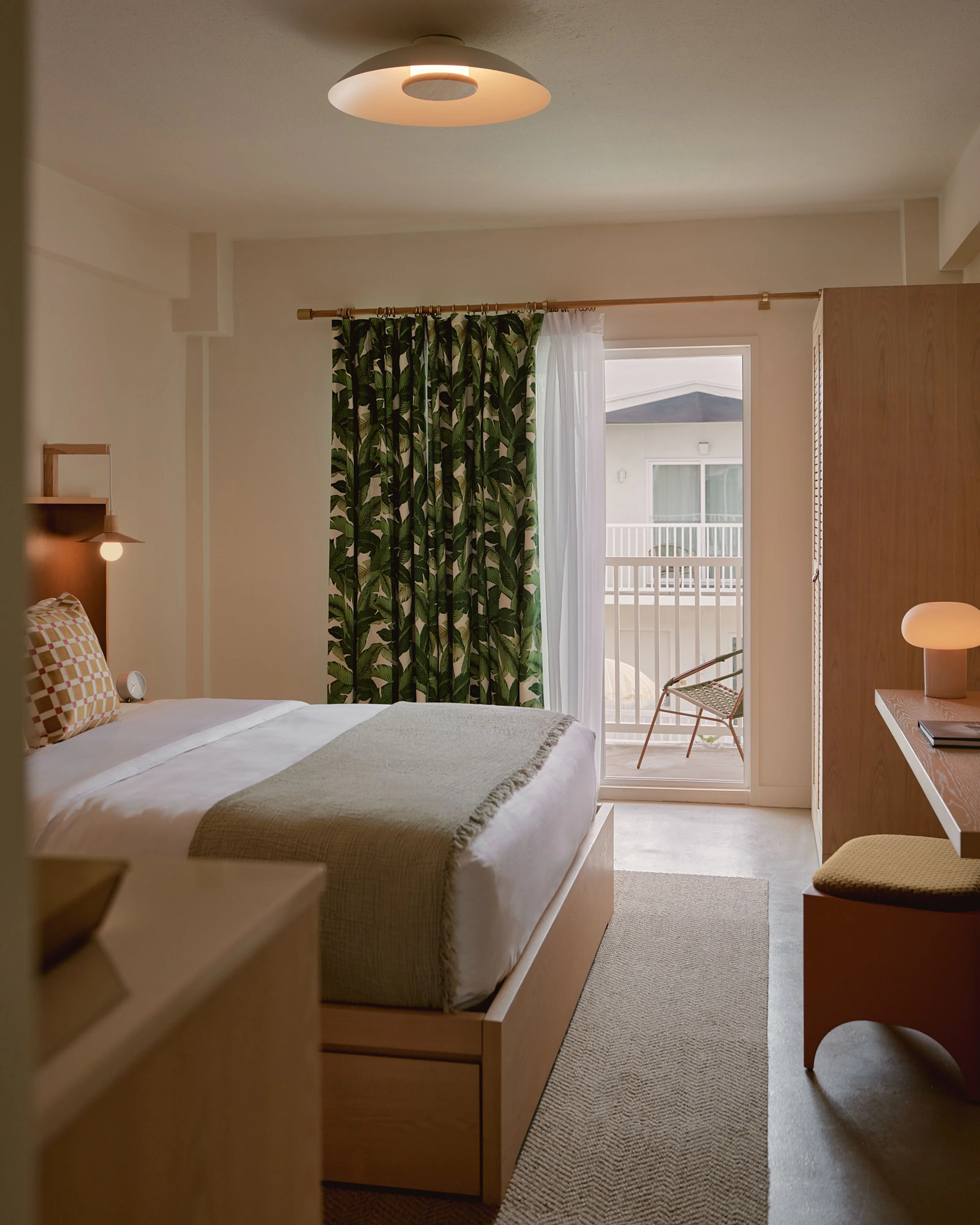 A serene modern hotel bedroom features warm wood furniture and neutral tones accented by bold botanical-patterned curtains in black and green; natural light floods through glass doors opening to a balcony, while a minimalist ceiling-mounted ring light and curated decor elements create a calm, contemporary atmosphere.