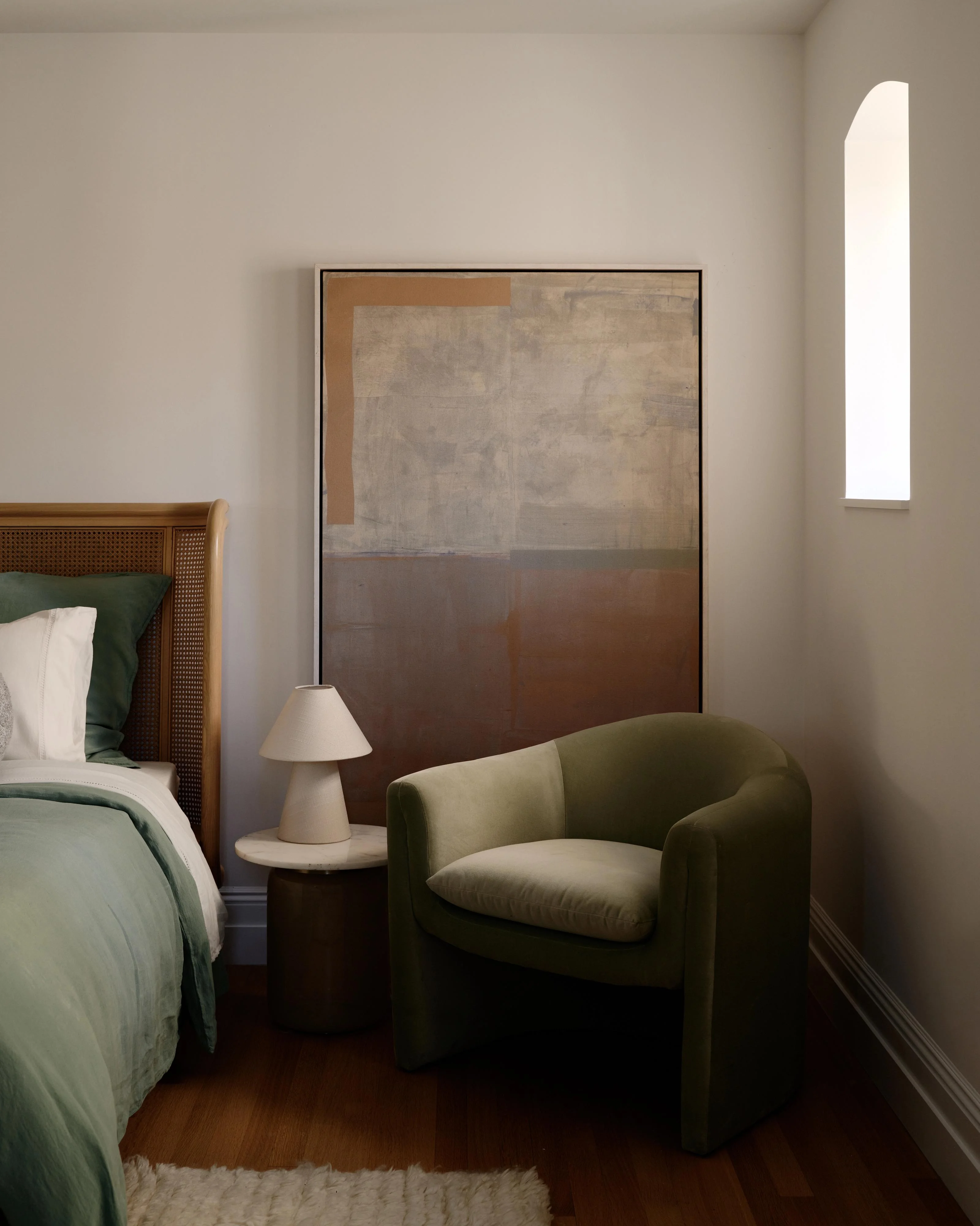 A serene, minimalist bedroom featuring a wooden-framed bed with rattan headboard and green bedding, paired with a sage-green upholstered armchair beside a small dark side table topped with a cream ceramic lamp. Soft natural light streams through a tall arched window, illuminating the cream walls and a large abstract artwork with muted earth tones.