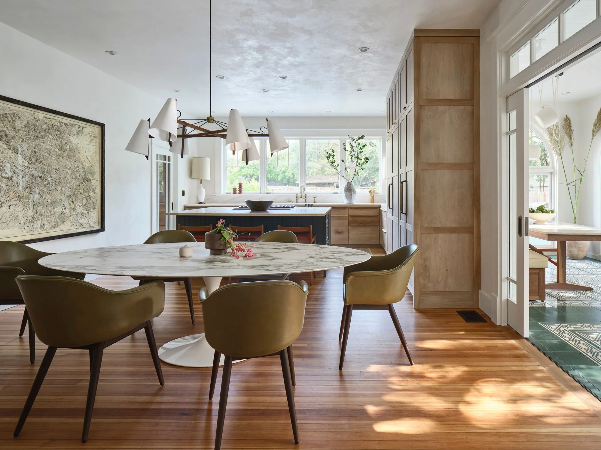 A bright, modern dining and kitchen space features a round marble-top table surrounded by olive-green upholstered chairs on warm hardwood floors, with a dark-island kitchen, pendant lighting, natural wood cabinetry, and expansive windows flooding the minimalist interior with natural light.
