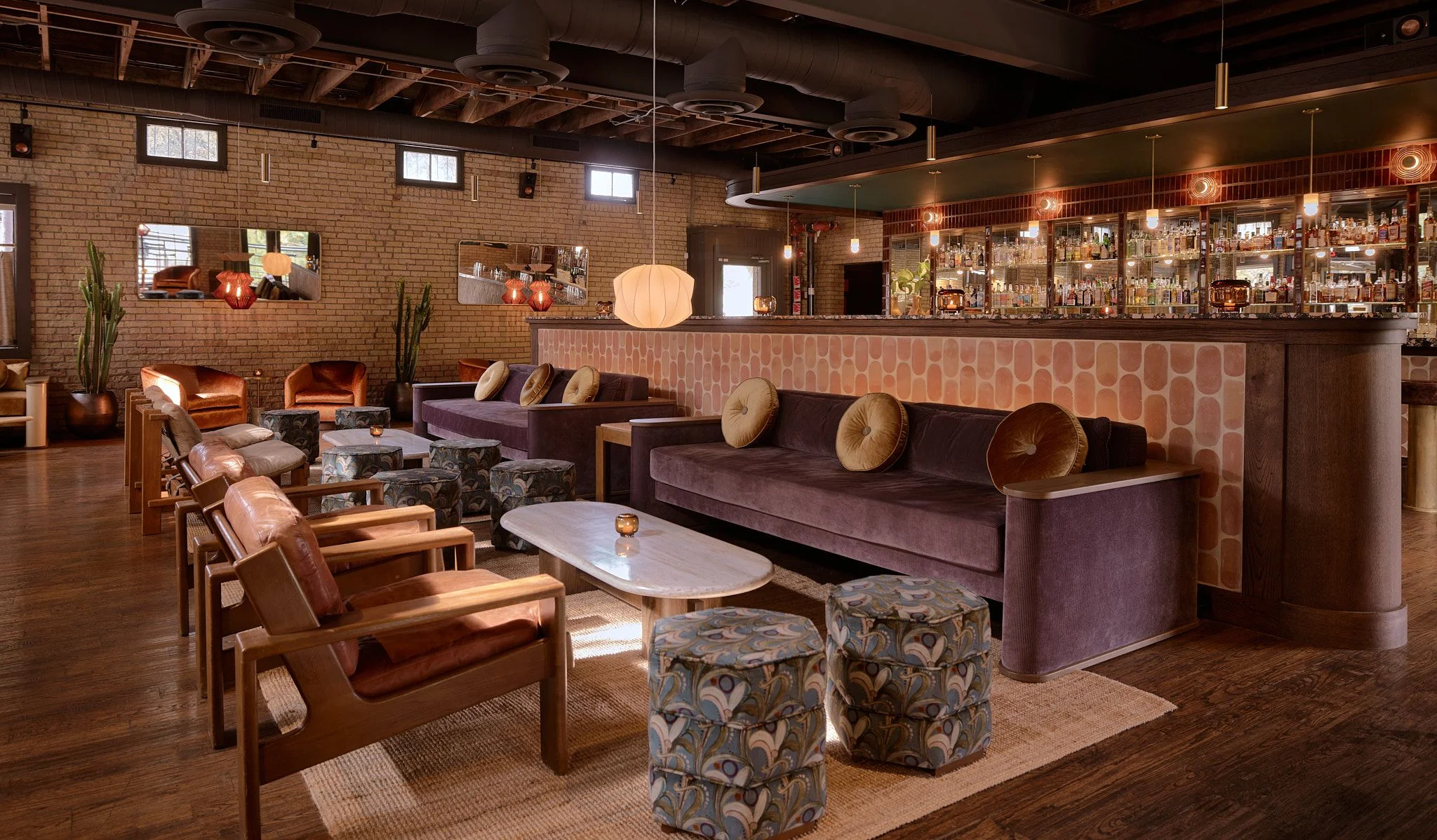 A sophisticated mid-century modern lounge features exposed brick walls, dark wooden beam ceilings, and warm ambient lighting from pendant fixtures and glowing orbs. The space is furnished with cognac leather chairs, plush purple velvet sofas, patterned ottoman seating, and marble-topped coffee tables, while a full bar with backlit shelving anchors the right side of the room.