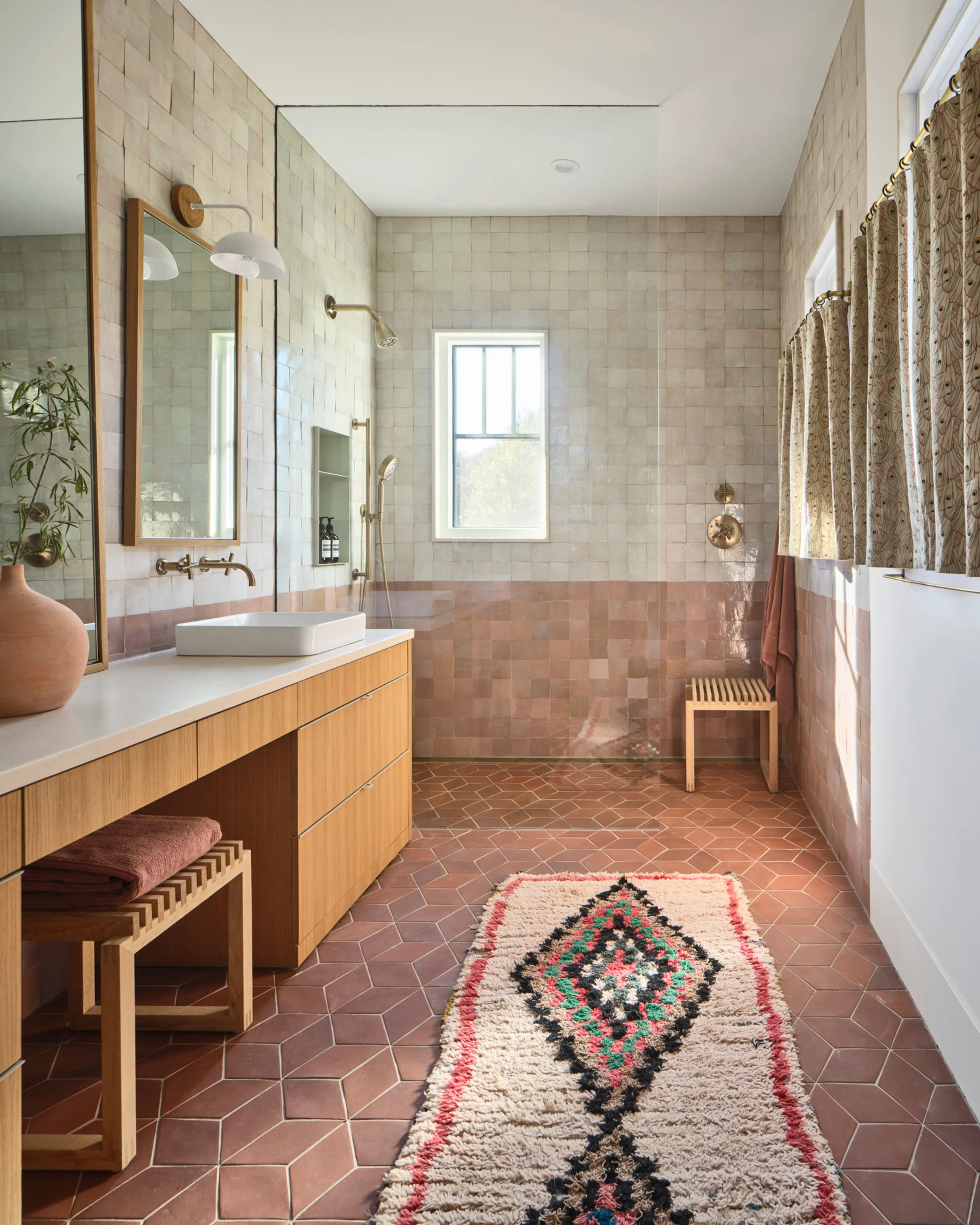 A spacious, light-filled bathroom features warm terracotta herringbone tile flooring, cream and dusty rose subway tile walls, and natural wood cabinetry with a white countertop and vessel sink. The room is accented with brass fixtures, a geometric patterned runner rug, a walk-in shower with glass enclosure, and soft natural light from a large window.
