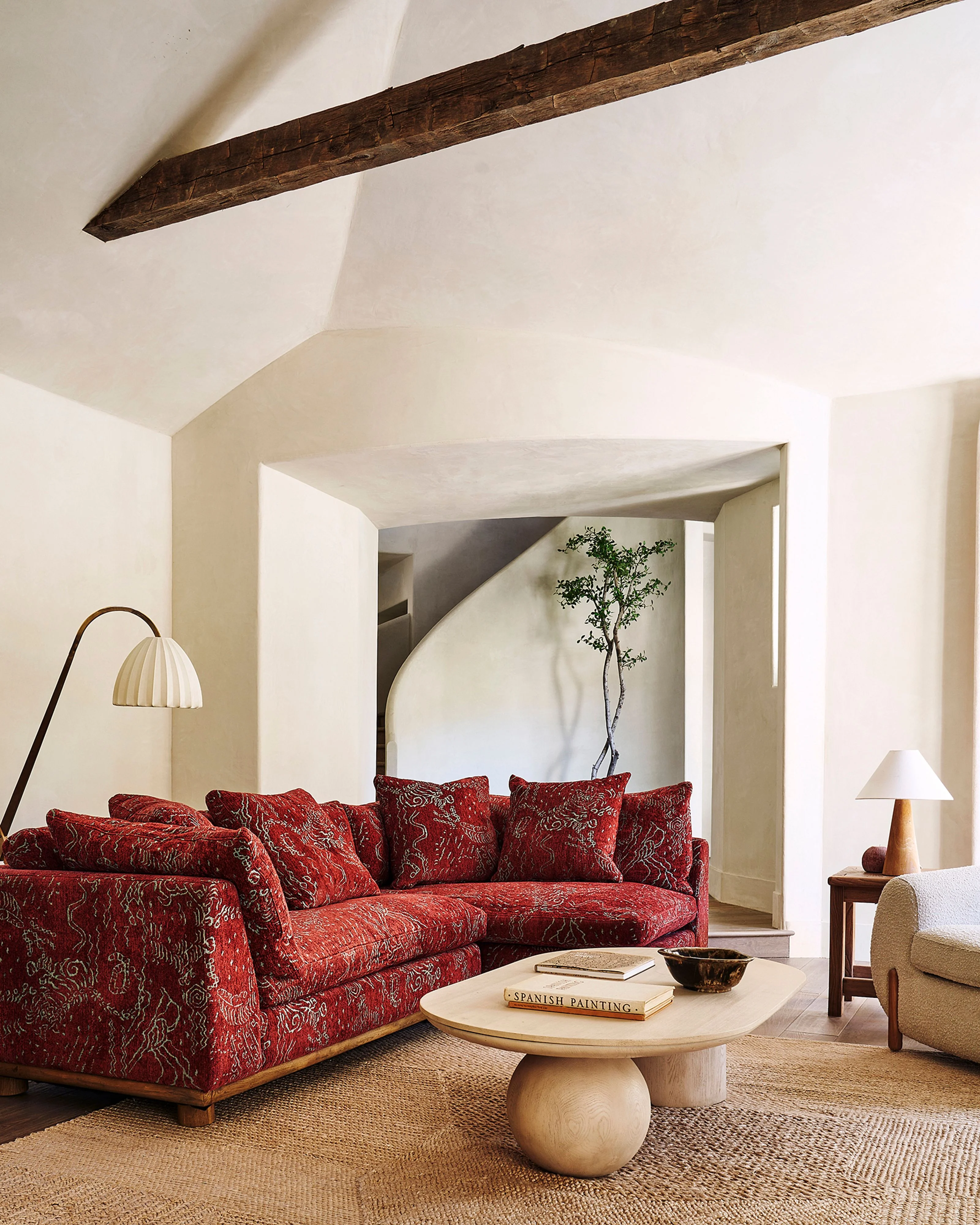 A bright, minimalist living room features a striking deep red patterned sectional sofa with wooden legs positioned beneath an exposed dark wooden beam against white walls and vaulted ceilings. The serene space is anchored by a sculptural round wooden coffee table, complemented by natural fiber rugs and a curved arc floor lamp.