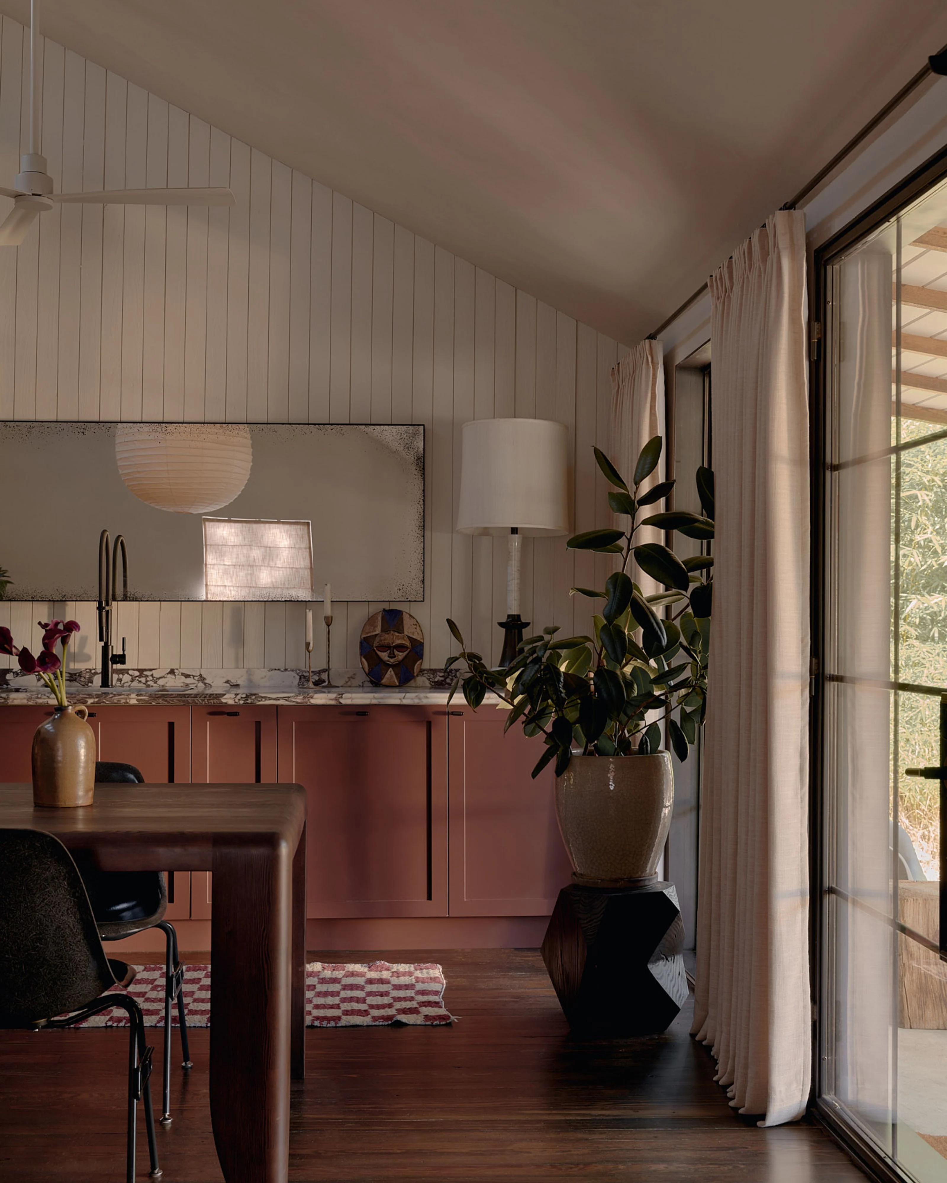 A modern, minimalist interior with warm terracotta cabinetry, white vertical shiplap walls, and dark wood flooring features a workspace with a round wooden desk, potted greenery, and a large mirror above the vanity; soft natural light streams through floor-to-ceiling glass doors draped with neutral linen curtains.