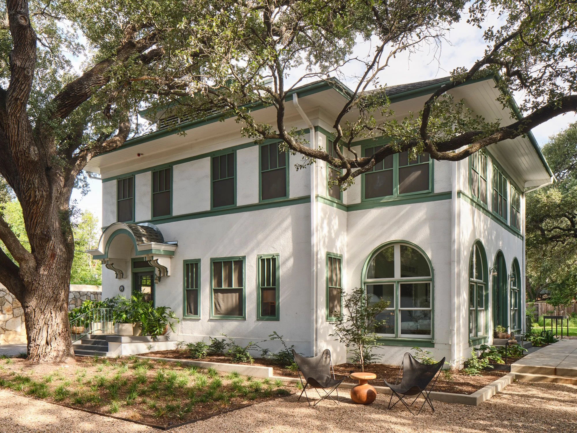 A modernist residential structure with crisp white walls and forest-green trim features clean geometric lines, large arched and rectangular windows, and a distinctive cantilevered roofline, nestled beneath mature oak trees with dappled natural light casting shadows across the contemporary landscape.