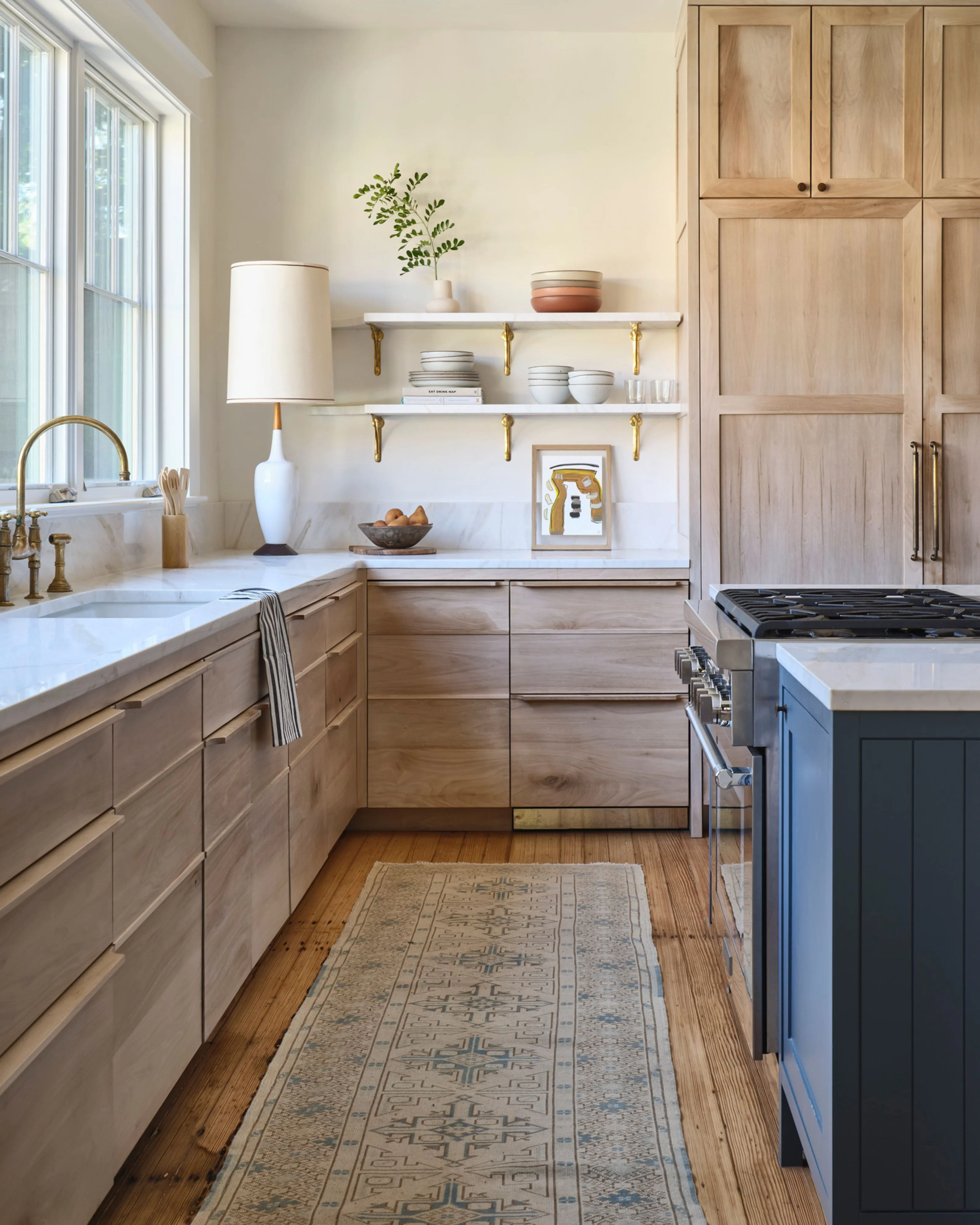 A bright, contemporary kitchen featuring natural wood cabinetry with marble countertops, white open shelving with brass brackets, and a mix of stainless steel and navy blue appliances, illuminated by abundant natural light from large windows and accented with warm brass hardware.