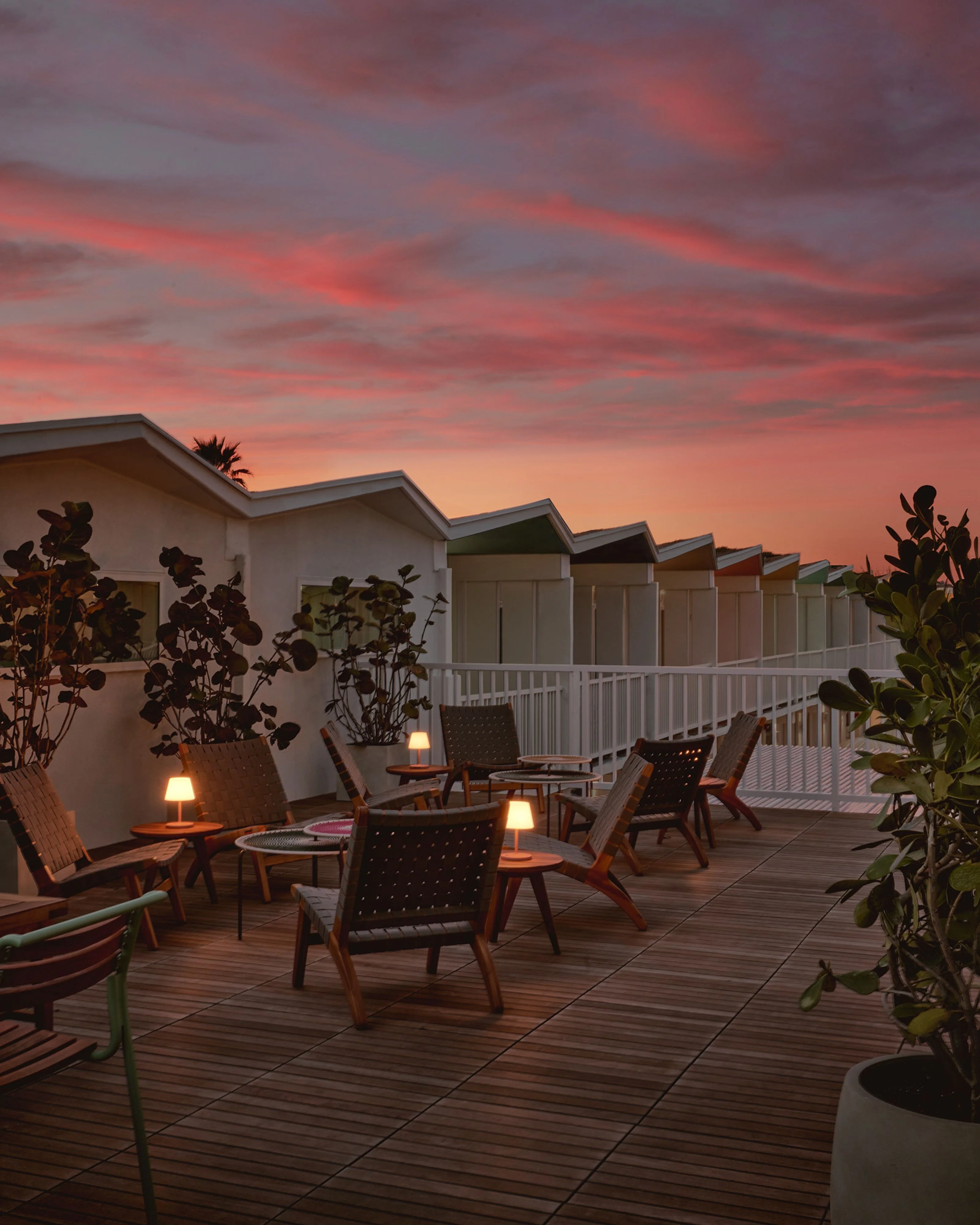 A modern beachfront promenade at sunset features a row of white beach cabins with pitched roofs and white railings, fronted by a wooden deck furnished with mid-century style lounge chairs and warm-glowing table lamps surrounded by potted tropical plants.