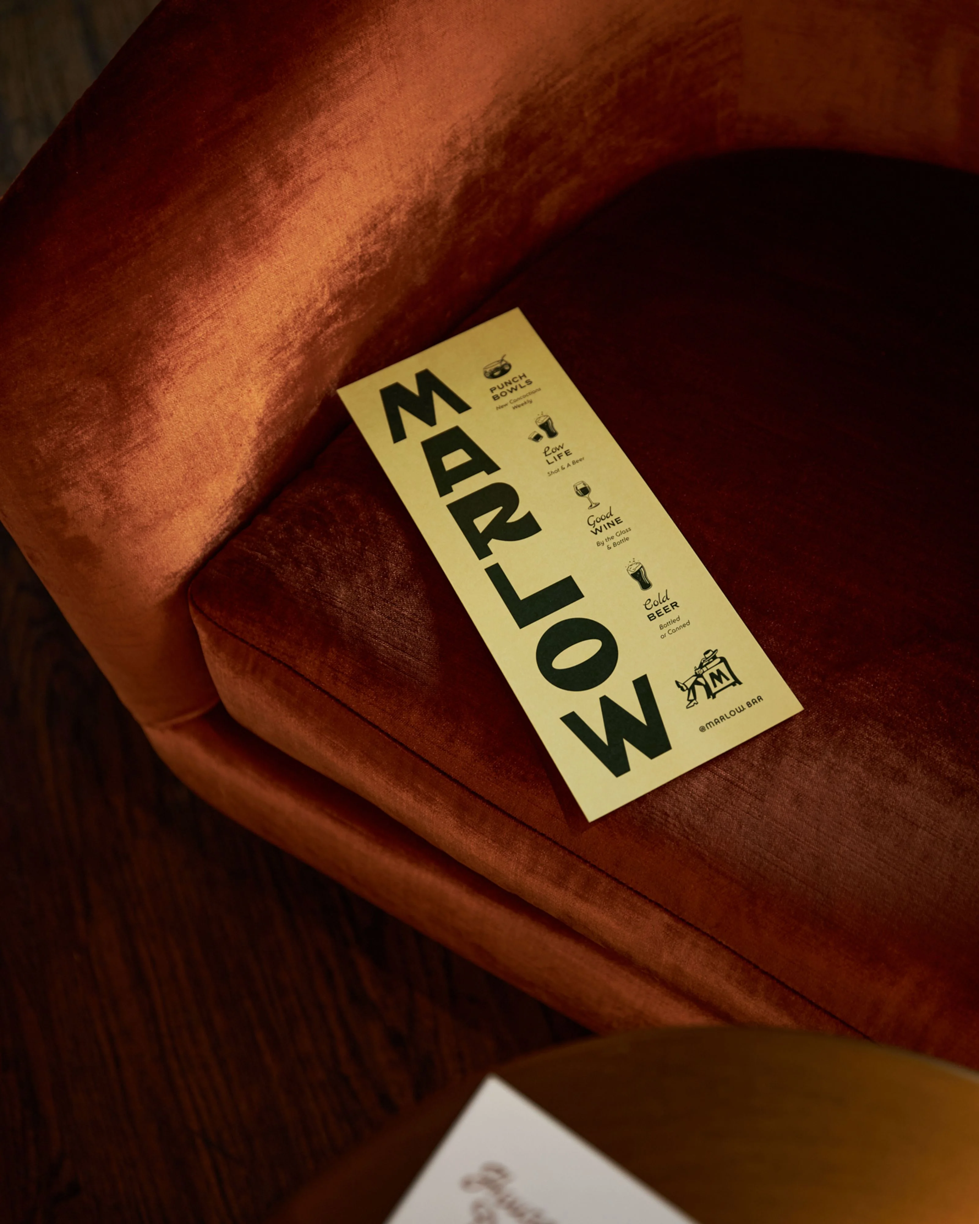 A cream-colored menu card labeled "MARLOW" in bold black typography rests on rich, rust-red velvet upholstery, creating a warm and luxurious aesthetic.