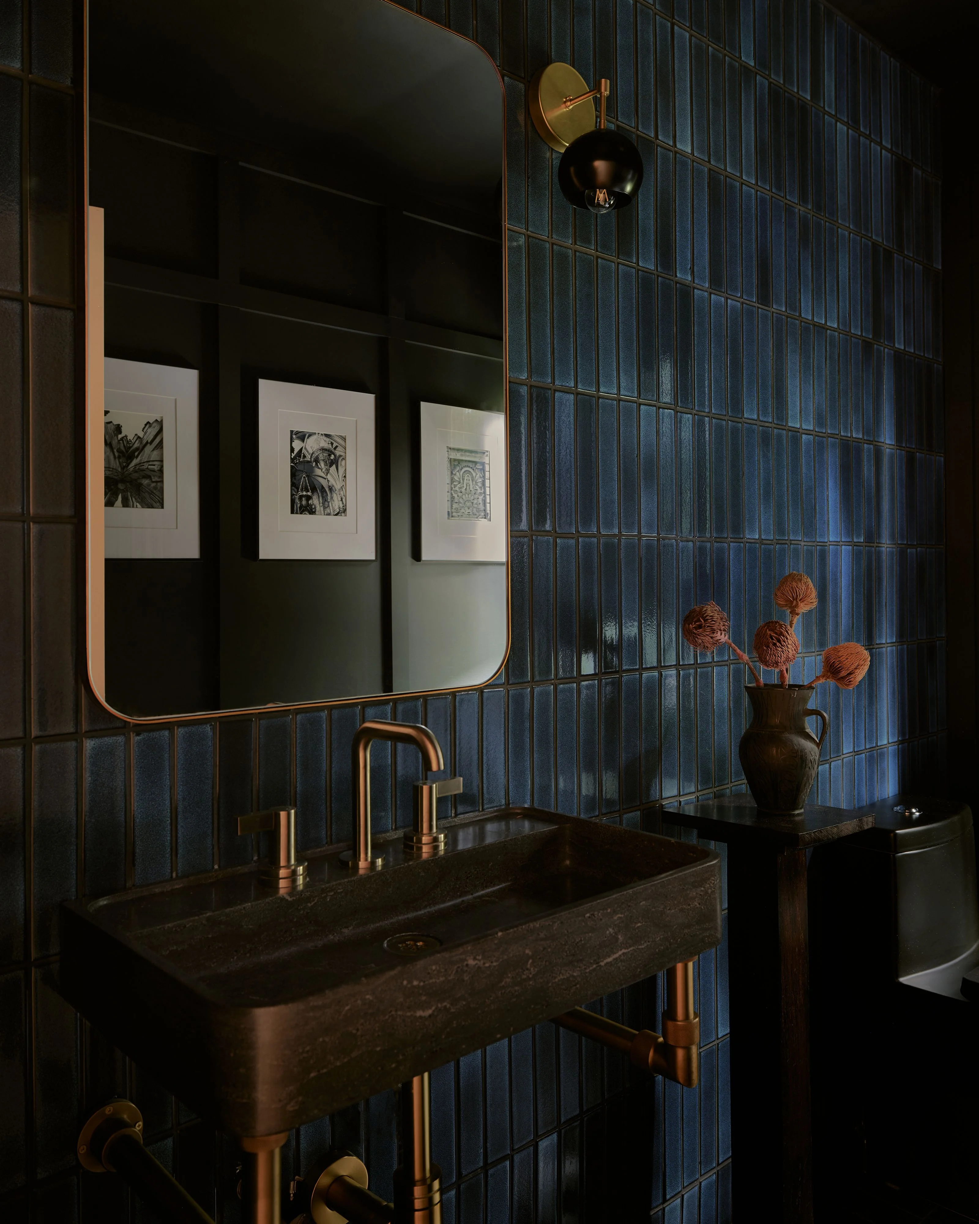 A moody modern bathroom features deep blue vertical tile walls, a dark stone sink with brass fixtures, and a rounded mirror with brass frame reflecting framed botanical artwork. Brass sconces and a black vase with dried flowers complete the sophisticated, minimalist aesthetic.