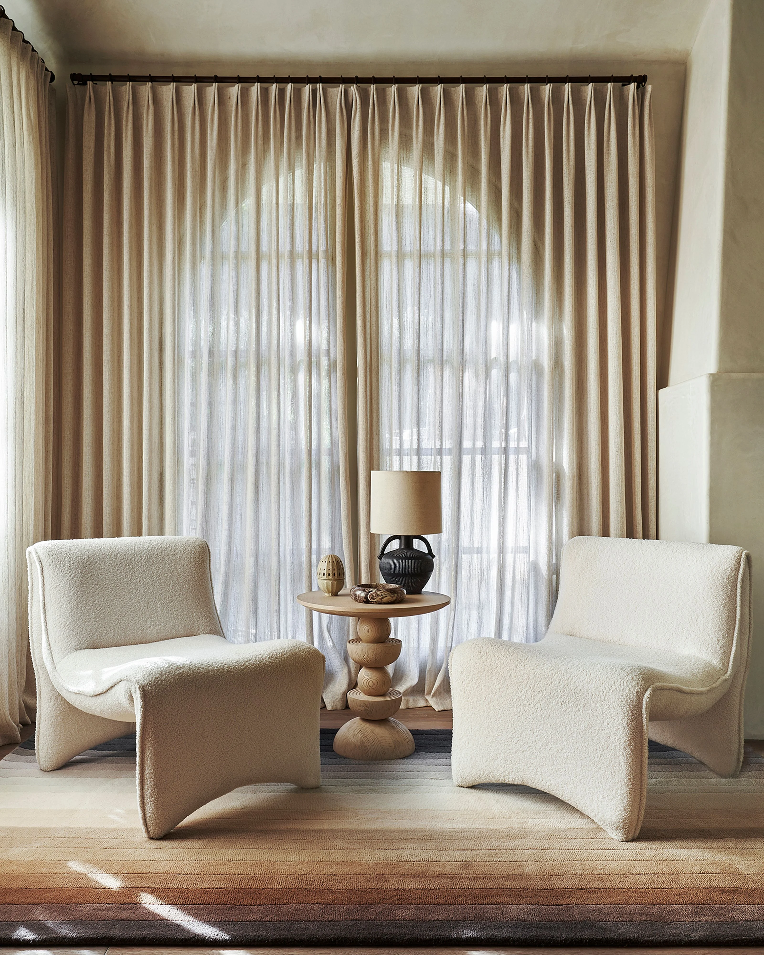 A serene, minimalist interior featuring two cream-colored sculptural armchairs facing each other across a wooden pedestal side table, all arranged on a neutral area rug in front of floor-to-ceiling windows dressed with layered pleated linen curtains in warm beige tones.