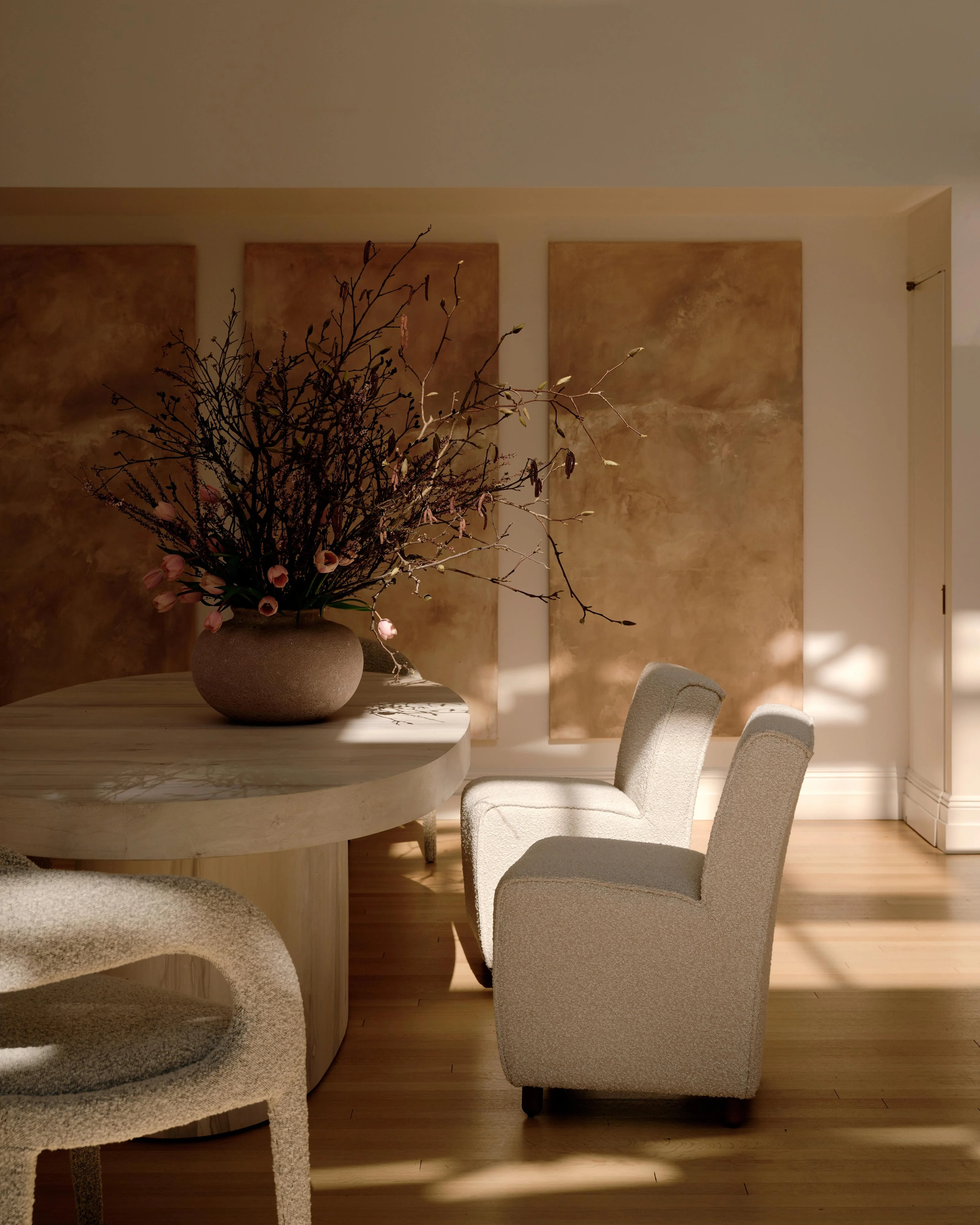 A serene, minimalist interior space bathed in warm natural light features a round light wood table topped with a sculptural arrangement of dried branches and pink blooms in a textured ceramic vessel, alongside soft cream-colored upholstered chairs and curved sculptural seating elements against warm earth-toned textured wall panels.