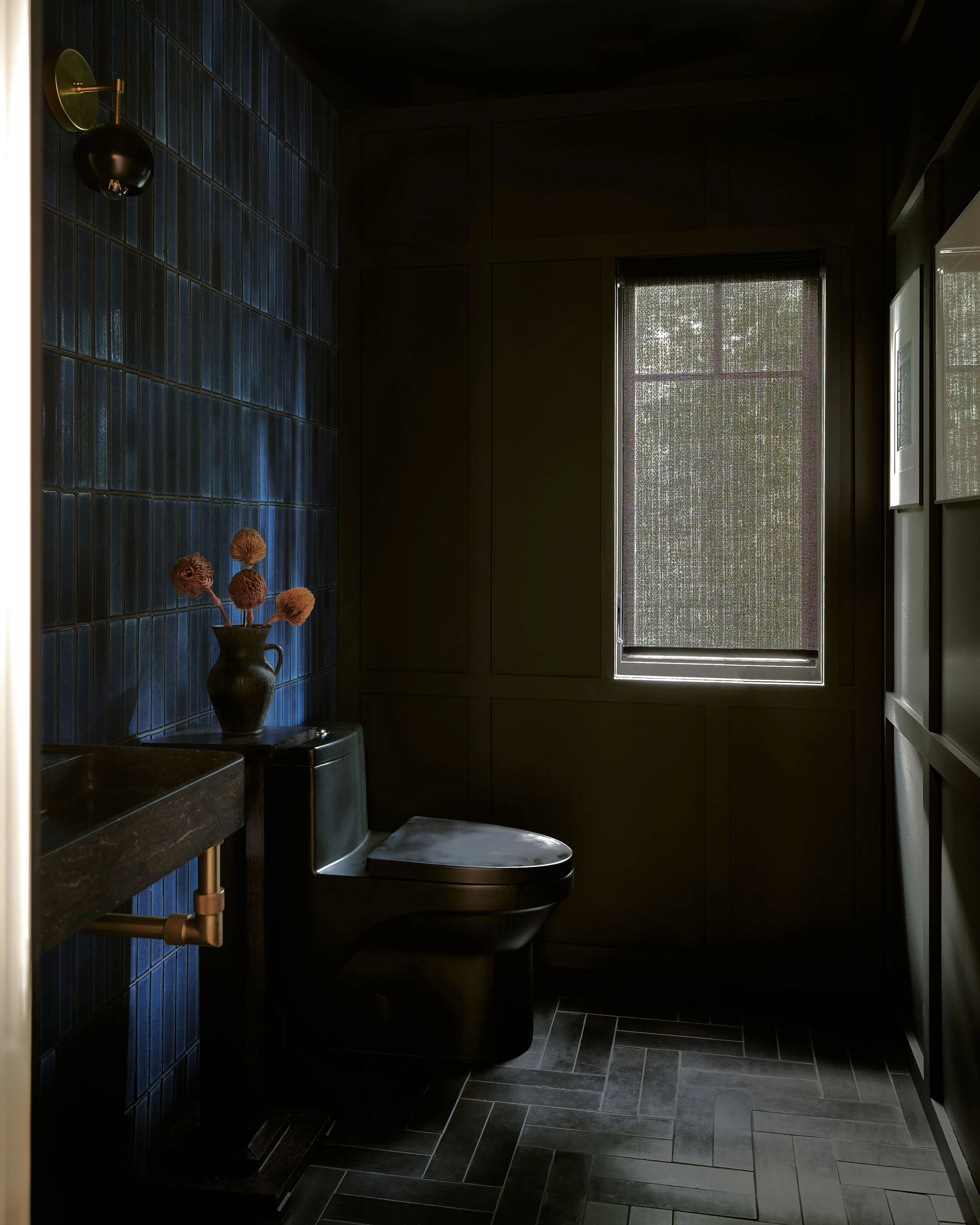 A moody, luxurious bathroom with deep black walls and dark herringbone tile flooring features a black toilet, a blue-tiled accent wall with brass fixtures, and a black vessel sink adorned with dried botanical stems. Natural light filters through textured window treatments, while decorative brass sconces and minimalist design elements create a sophisticated, contemporary aesthetic.