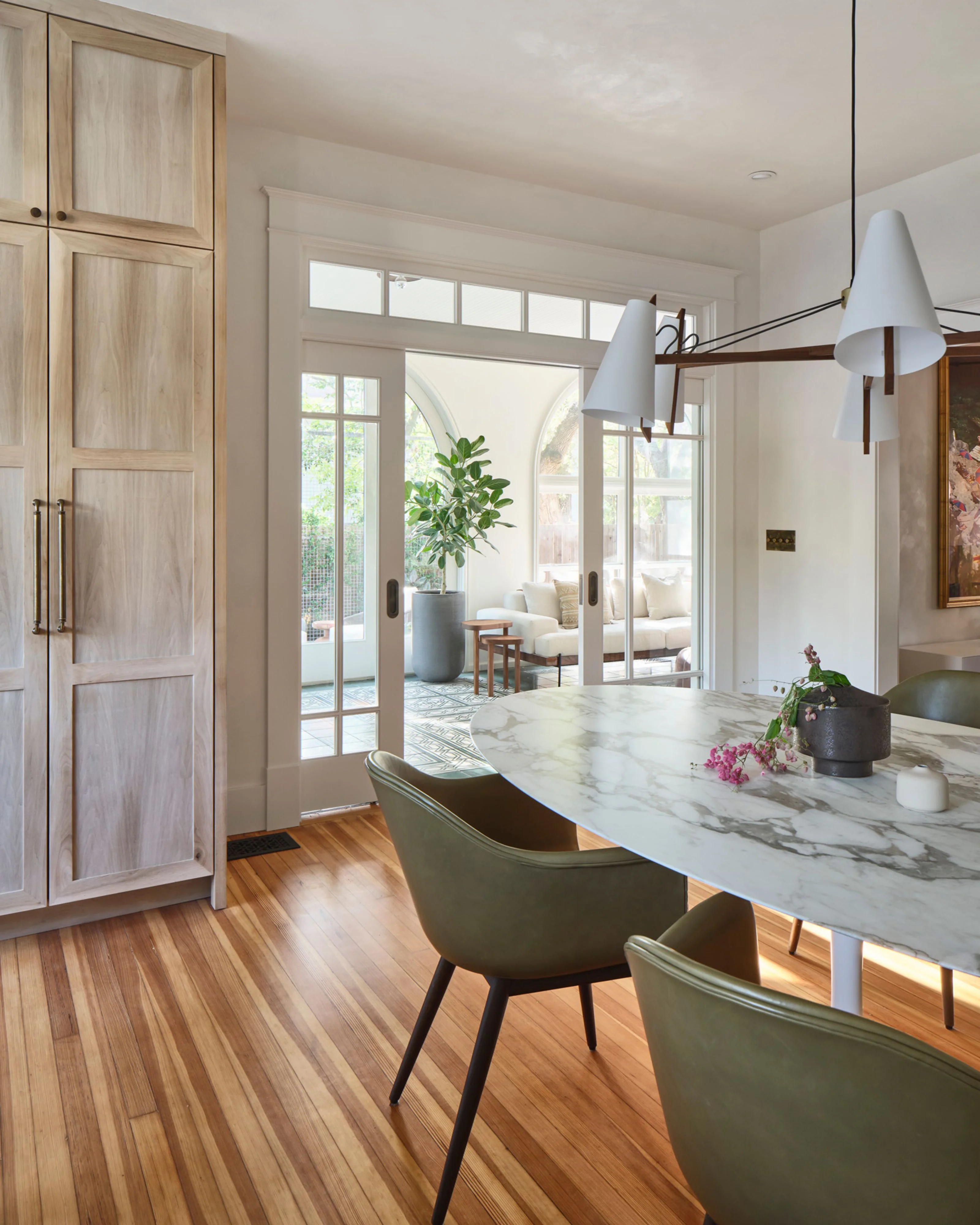 A bright, contemporary kitchen features a marble-topped island with sage green upholstered seating, warm wood flooring, and light wood cabinetry, while large glass doors open to a sunlit patio. Pendant lighting with white shades hangs above the island, creating an airy, modern aesthetic.