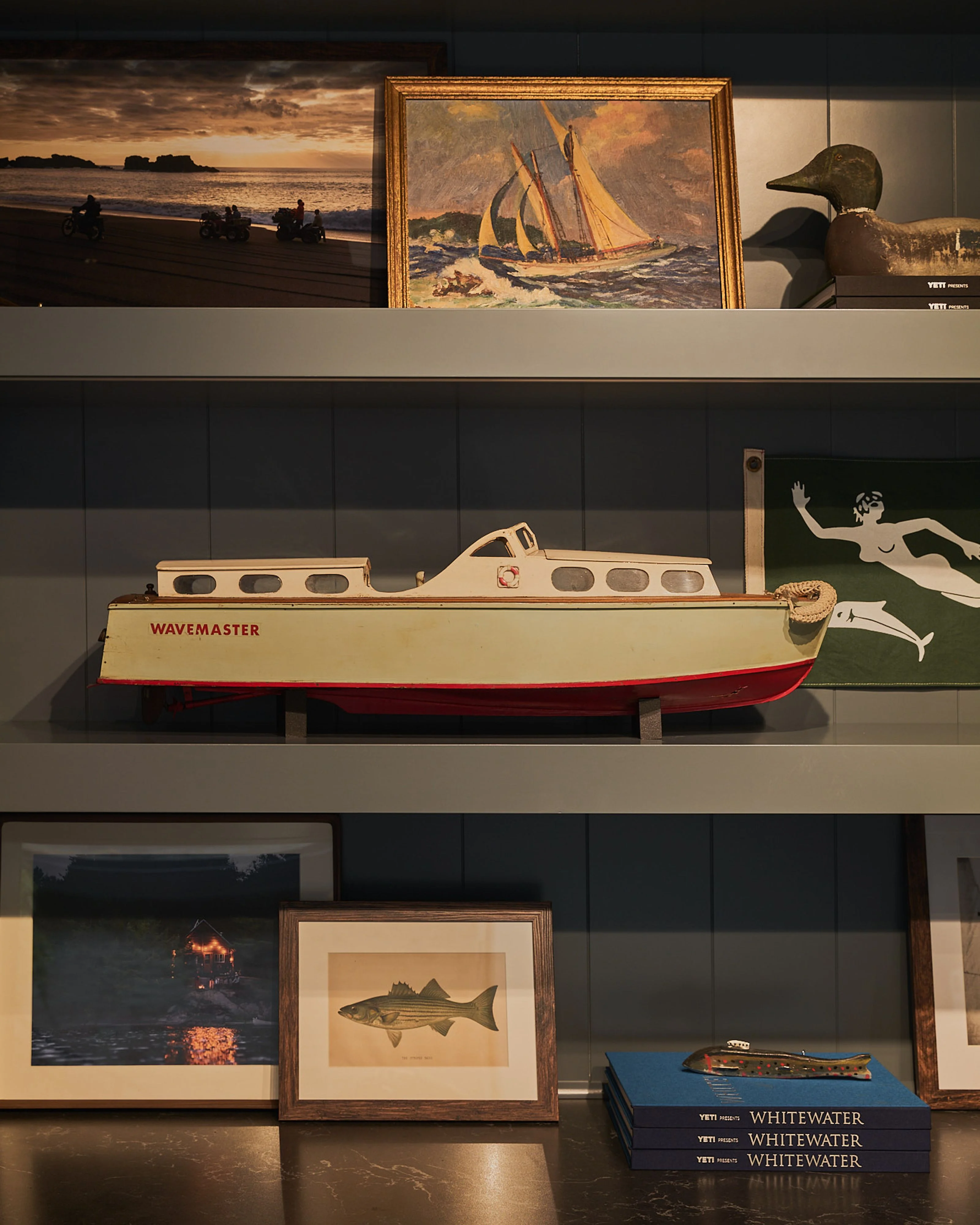 A museum display shelf features a nautical and maritime-themed collection arranged across three tiers against dark paneled walls, including a cream and red wooden boat model, framed paintings of sailing ships and seascapes, a vintage fish illustration, decorative duck decoys, and stacked books, creating a cohesive coastal aesthetic with warm gallery lighting.