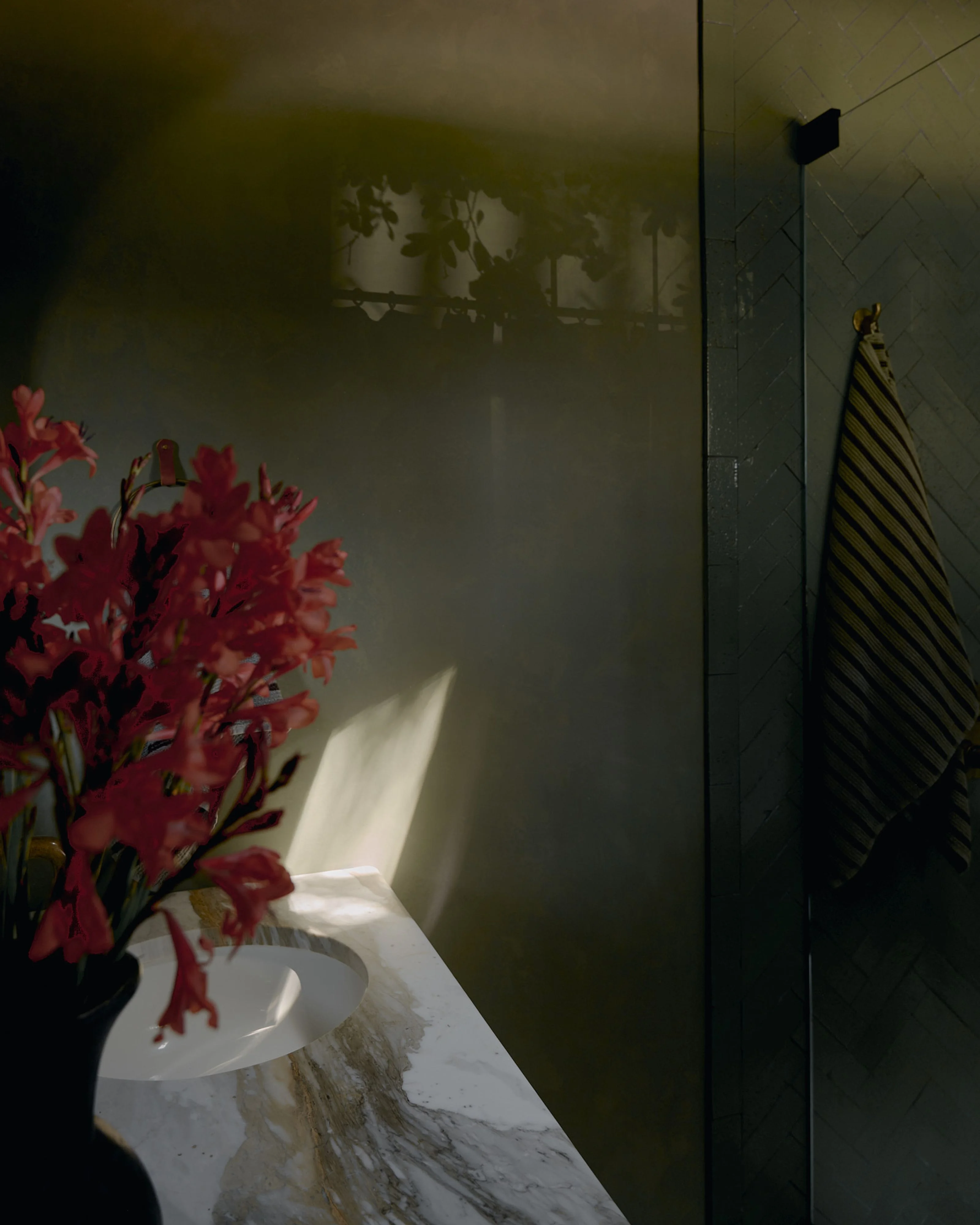 A minimalist interior space featuring a marble windowsill with a white vase of vibrant red flowers illuminated by soft natural light, while a dark herringbone-patterned radiator and muted olive-green walls create a moody, contemporary aesthetic.