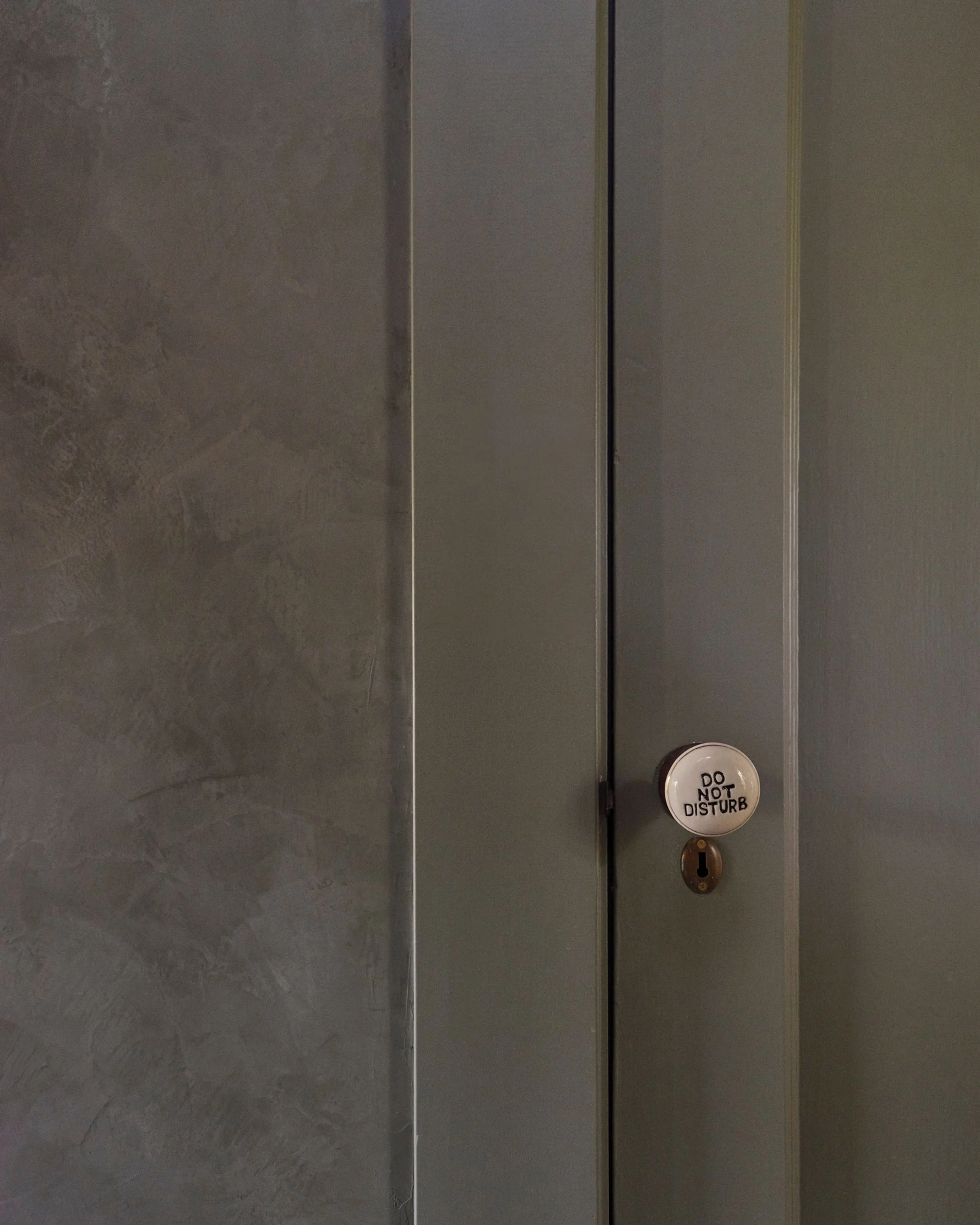 A minimalist interior view of a neutral-toned door with vertical panel details and a brass keyhole, featuring a small circular sign mounted in the center. The subdued gray and beige palette and clean lines create a contemporary, understated aesthetic.