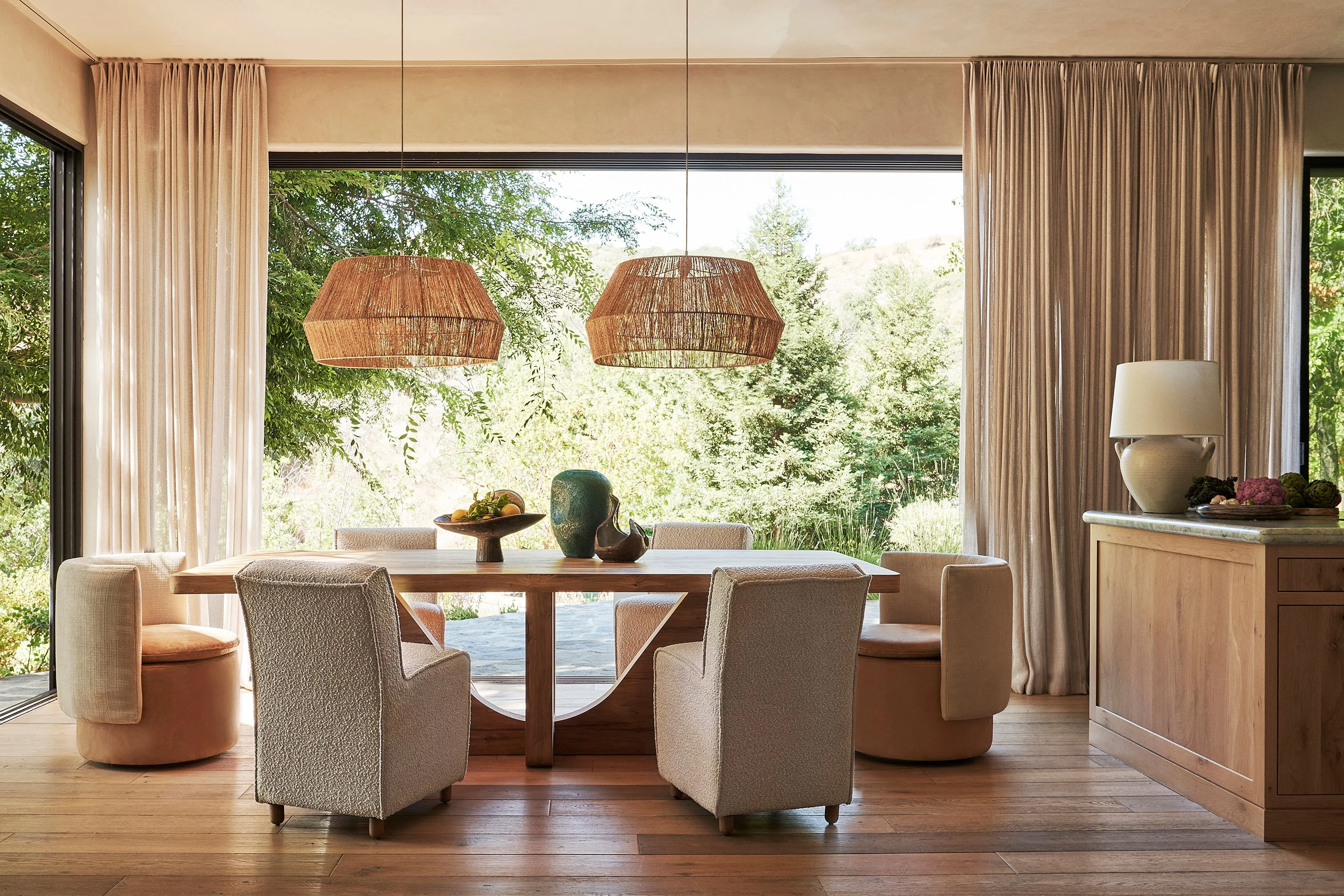 A serene dining room with warm neutral tones features a wooden table surrounded by upholstered chairs, illuminated by two woven rattan pendant lights; floor-to-ceiling windows with taupe linen curtains frame expansive views of lush green forest and natural landscape, while warm wood flooring and natural light create an open, tranquil atmosphere.