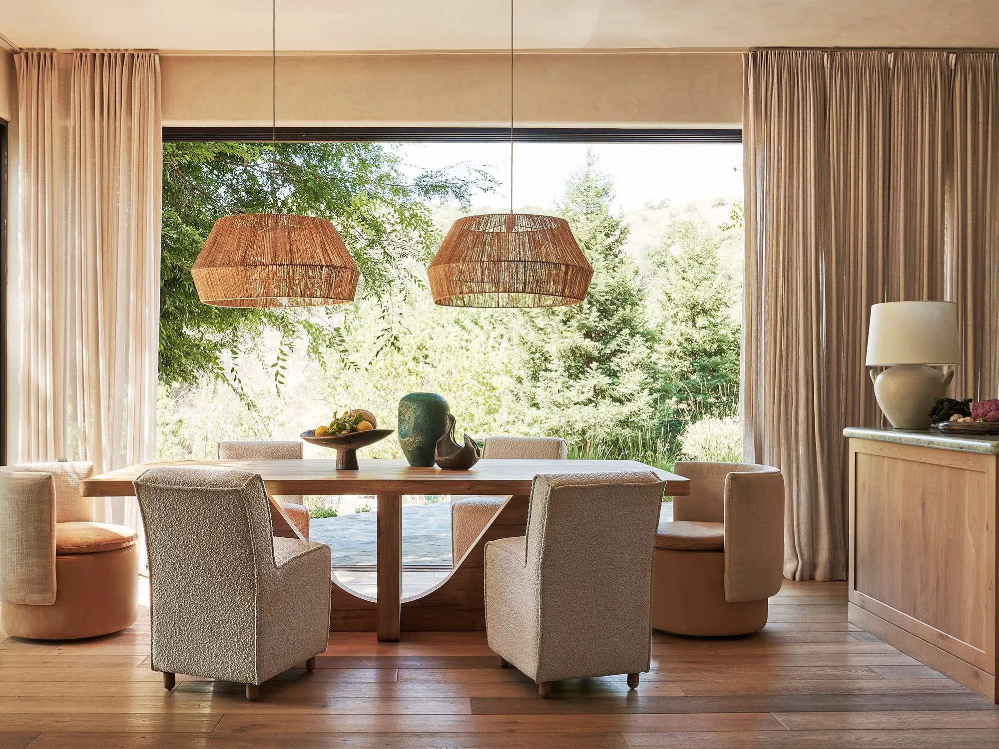 A serene dining room with warm neutral tones features a wooden table surrounded by upholstered chairs, illuminated by two woven rattan pendant lights; floor-to-ceiling windows with taupe linen curtains frame expansive views of lush green forest and natural landscape, while warm wood flooring and natural light create an open, tranquil atmosphere.