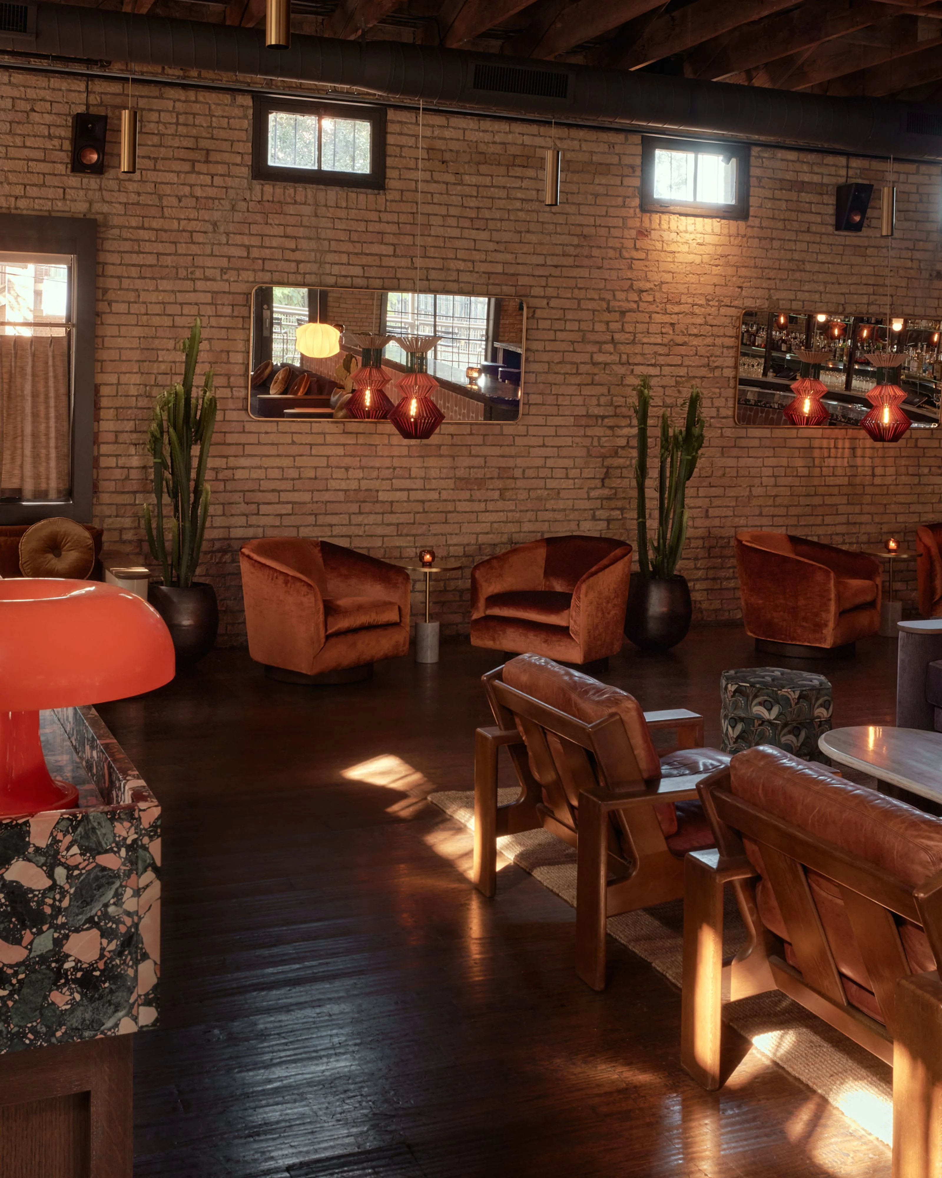 A mid-century modern lounge space features warm terracotta brick walls, exposed wooden ceiling beams, and dark hardwood flooring illuminated by natural light from large windows. The room is furnished with rust-colored velvet armchairs, a red accent table, tall green cacti, and copper pendant lights.