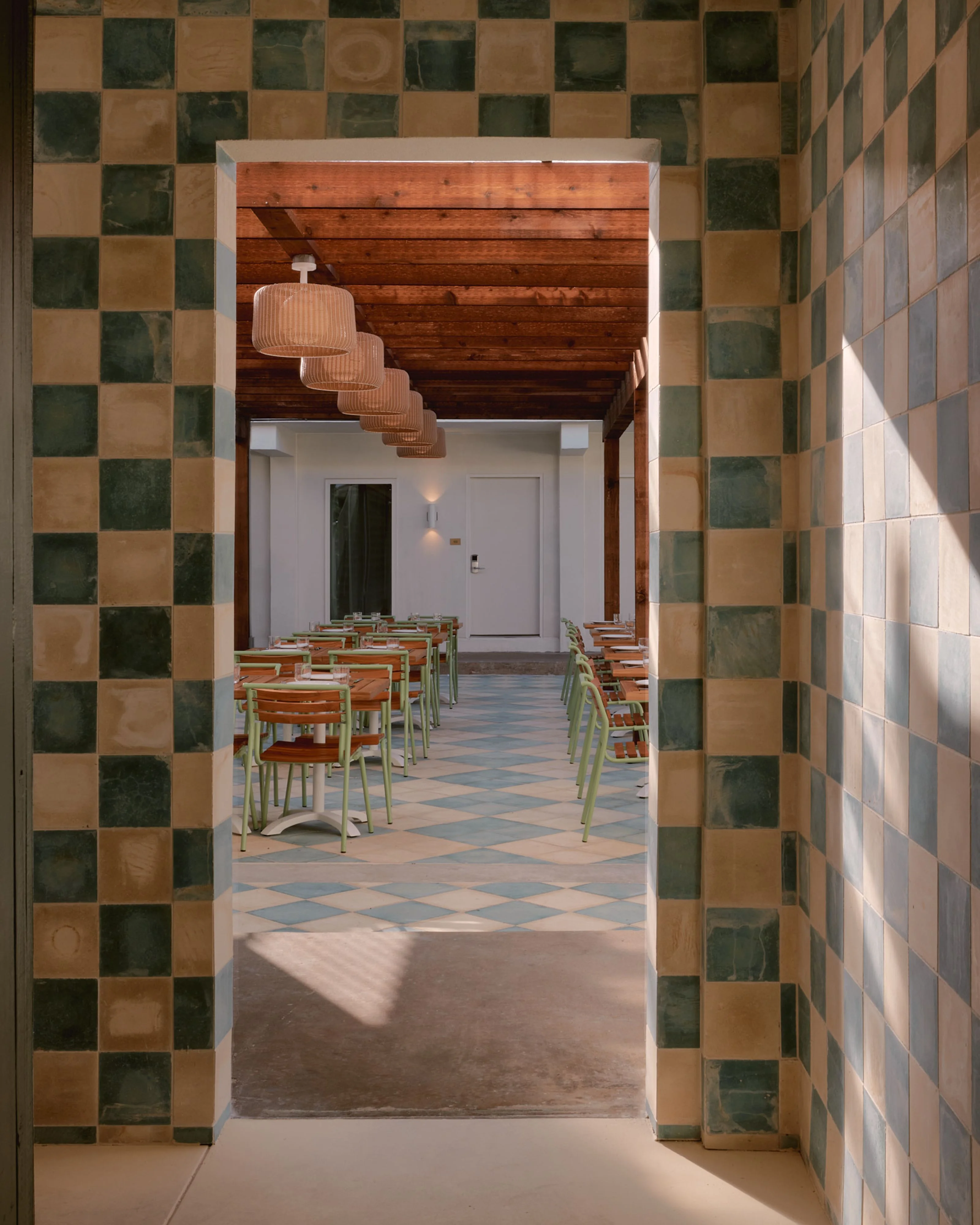 A modern dining space viewed through a checkerboard-patterned tile doorway, featuring wooden chairs arranged at tables beneath a warm wood ceiling with suspended ribbed pendant lights; the room is flooded with natural daylight and accented by geometric diamond-patterned floor tiles in muted blue and tan tones.