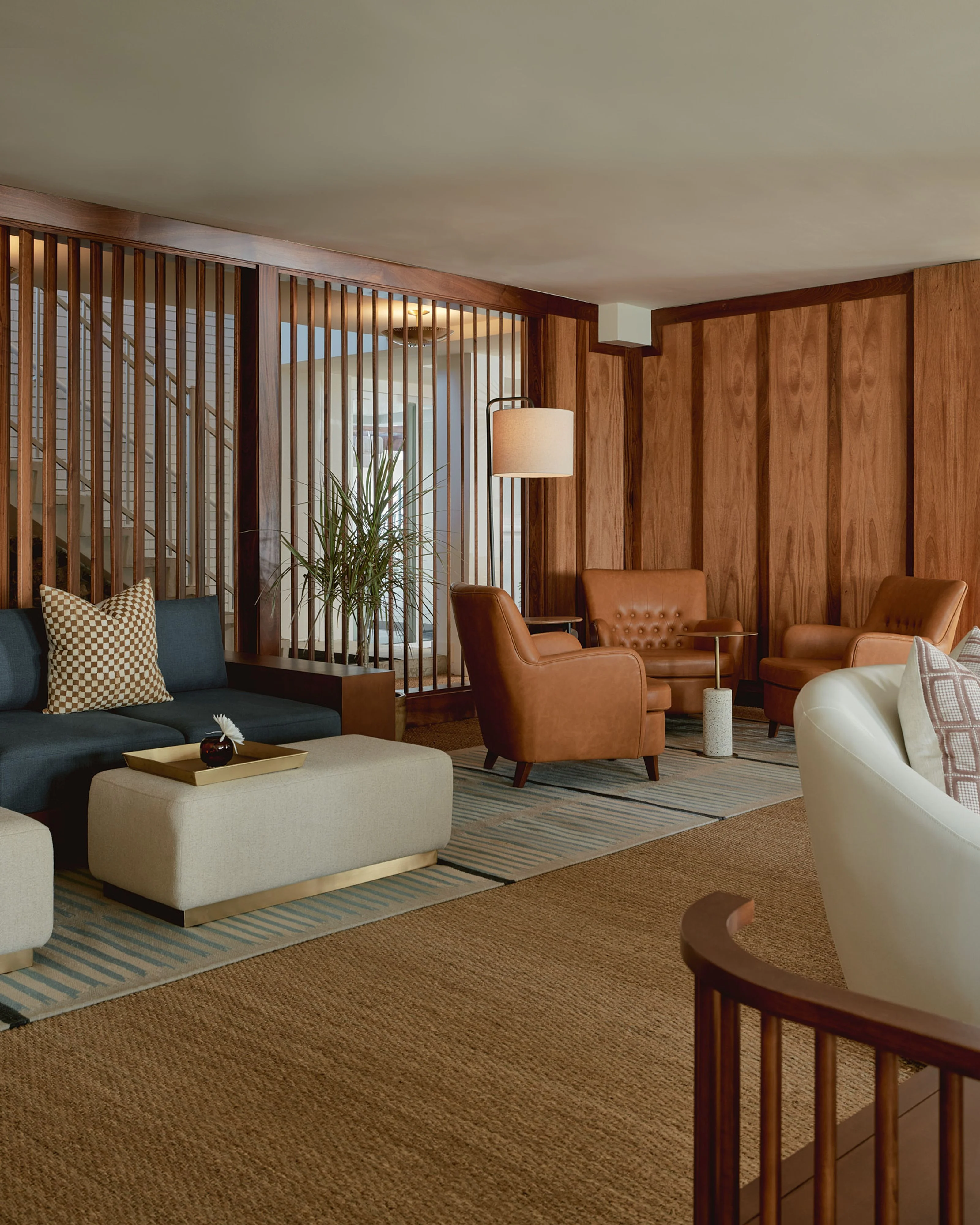 A mid-century modern lounge space featuring rich walnut wood paneling, warm cognac leather armchairs, and cream-colored upholstered ottomans arranged on a striped area rug, with vertical wood slat room dividers filtering natural light and creating intimate seating zones.