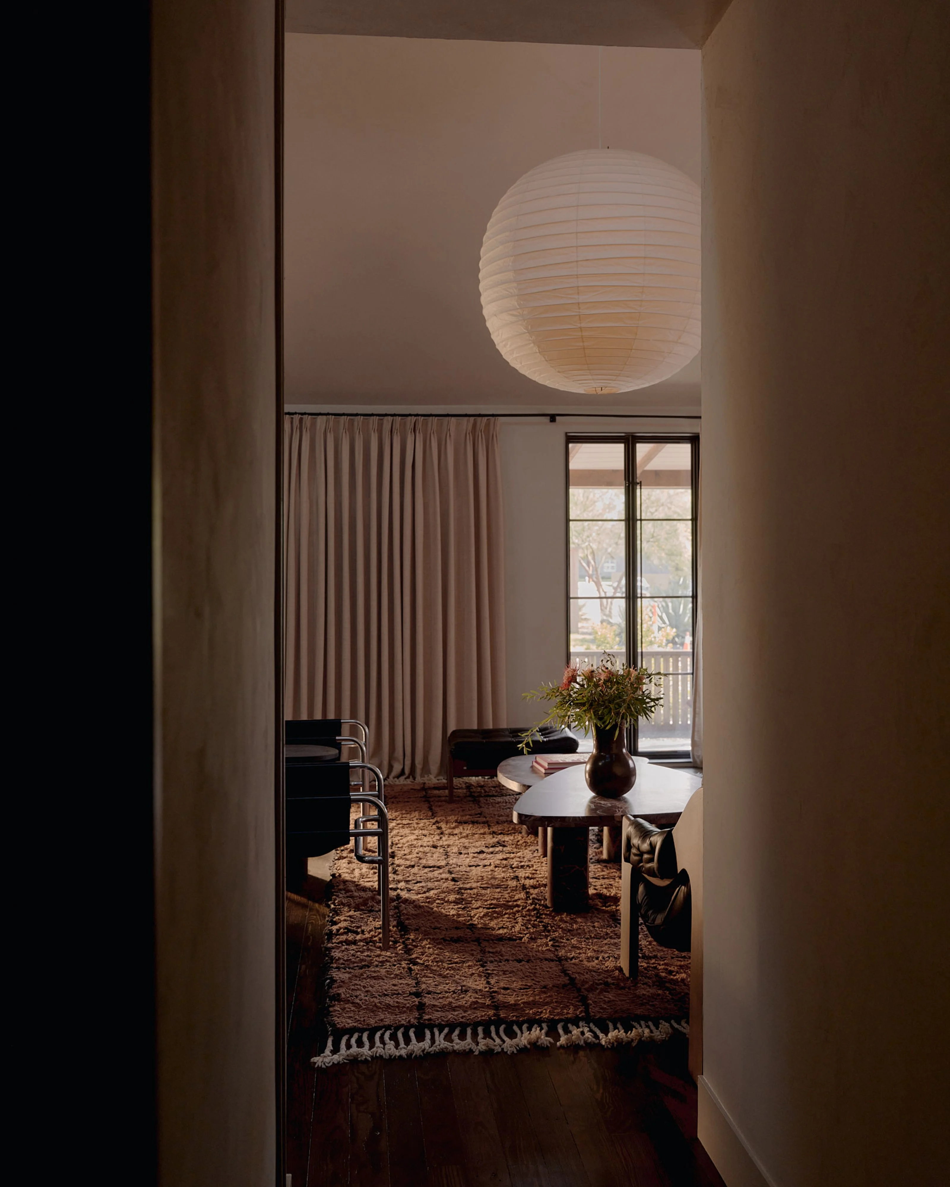 A serene, minimalist interior viewed through an open doorway features warm neutral tones, cream-colored walls, and a striking white paper lantern pendant light suspended from the ceiling. The space includes a small round black table with fresh flowers, mid-century modern seating, textured terracotta area rug, and floor-to-ceiling curtains.