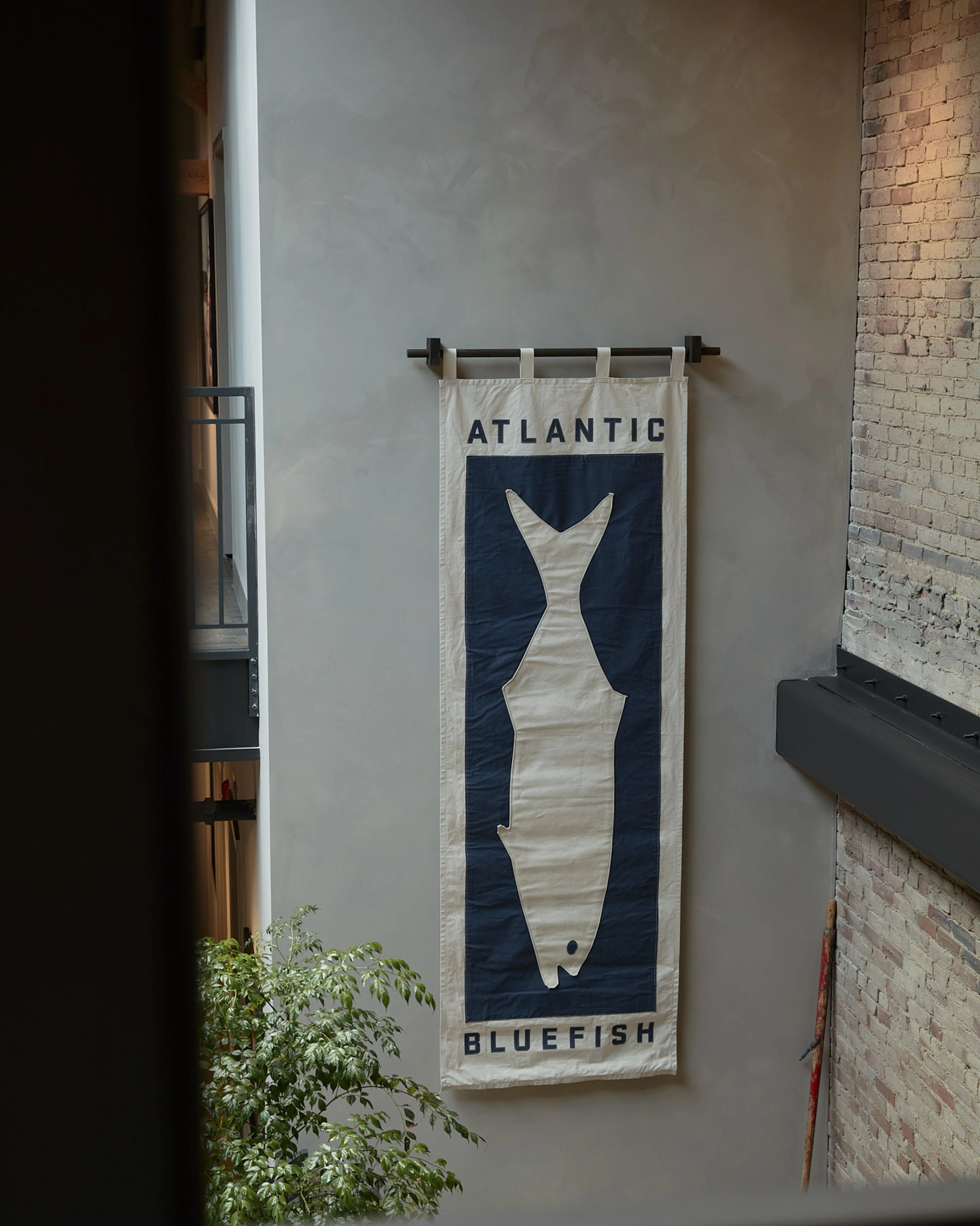 A minimalist industrial space features an exposed red brick wall on the right and a neutral gray wall displaying a vintage-style banner reading "ATLANTIC BLUEFISH" with a white fish silhouette on dark navy fabric, hung from a black metal rod.