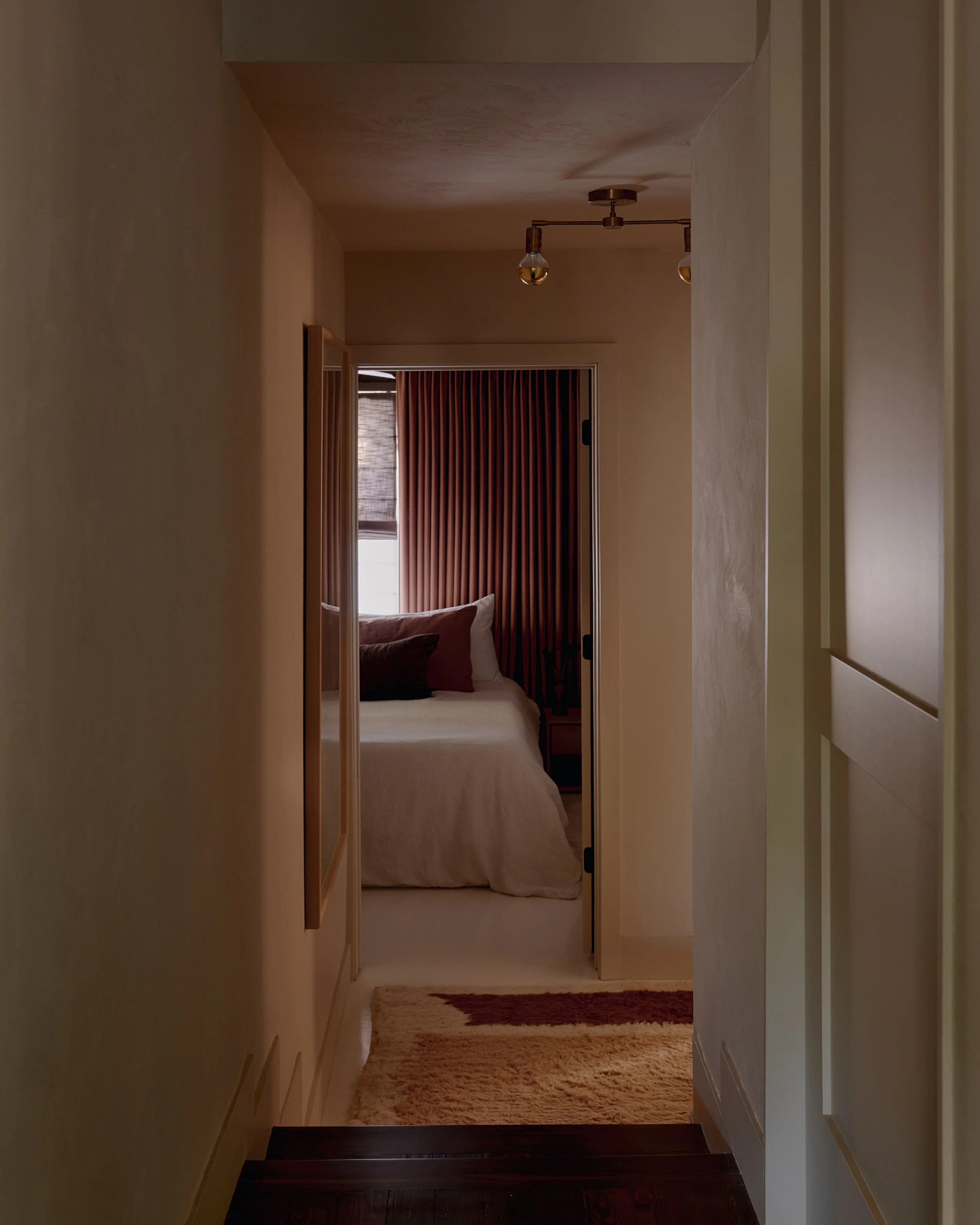 A minimalist hallway with cream-colored walls and dark wood flooring leads to a serene bedroom featuring deep burgundy vertical curtains, a bed with coordinating pillows, and warm ambient lighting. The clean lines, neutral palette, and careful use of warm tones create a calm, contemporary aesthetic.