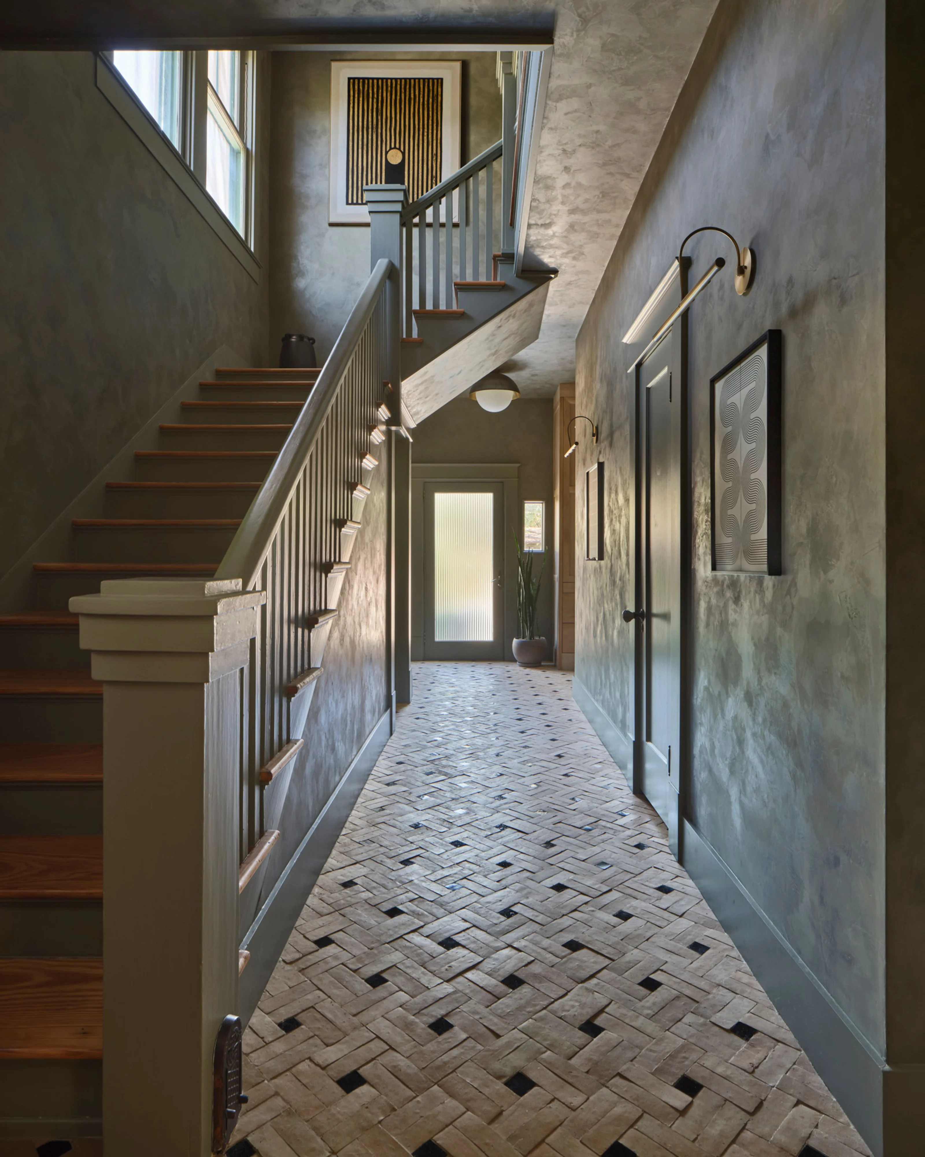 A contemporary hallway with textured plaster walls and herringbone-patterned tile flooring features a wooden staircase with metal railings on the left, brass hardware accents, and natural light streaming through windows, creating a modern yet warm transitional space.