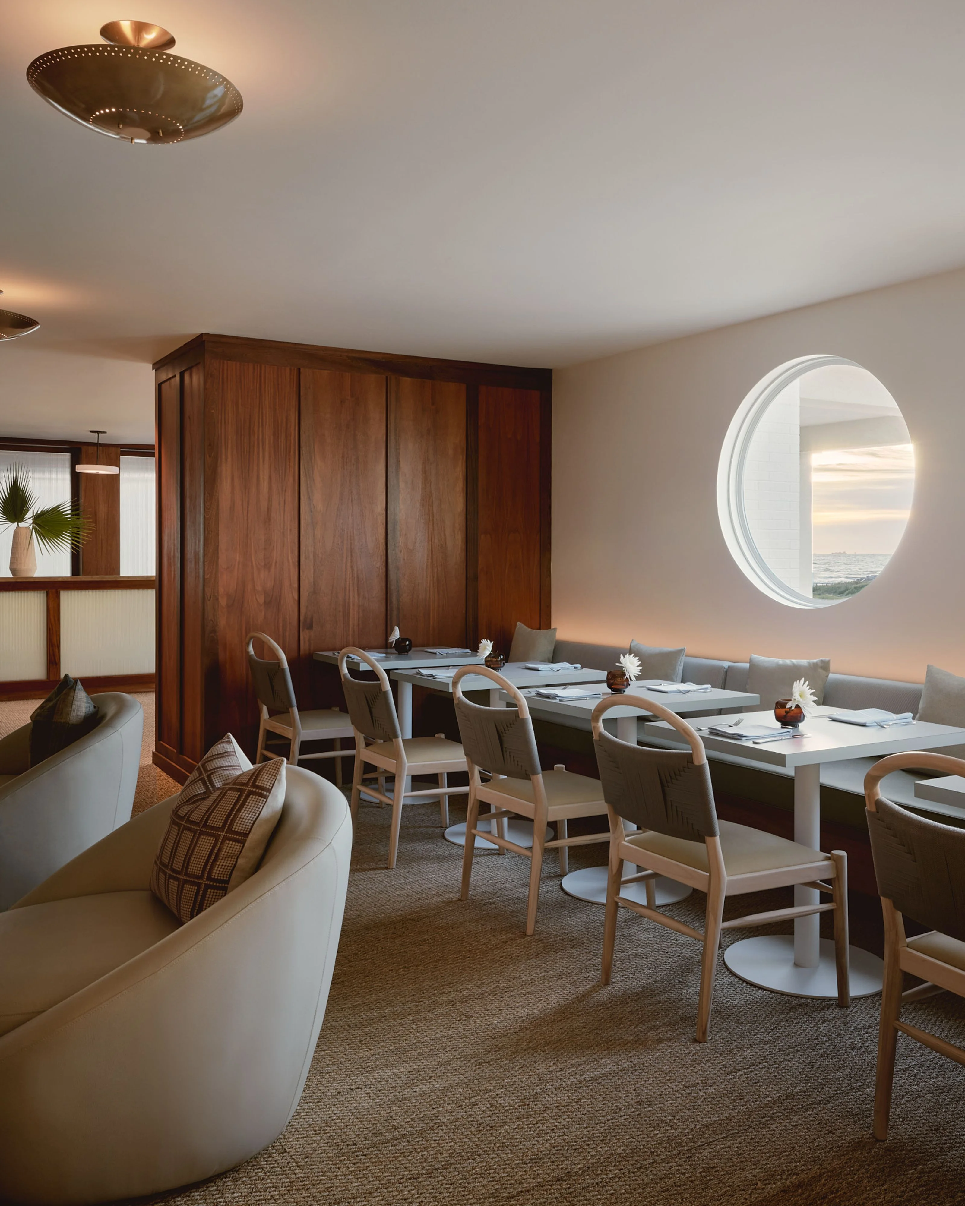 A modern dining space with warm, neutral tones features mid-century wooden dining tables and light wood chairs, complemented by a cream curved sofa and rich wood cabinetry; natural light streams through a large circular porthole window offering ocean views, while brass ceiling fixtures and neutral carpeting complete the sophisticated, maritime-inspired interior.