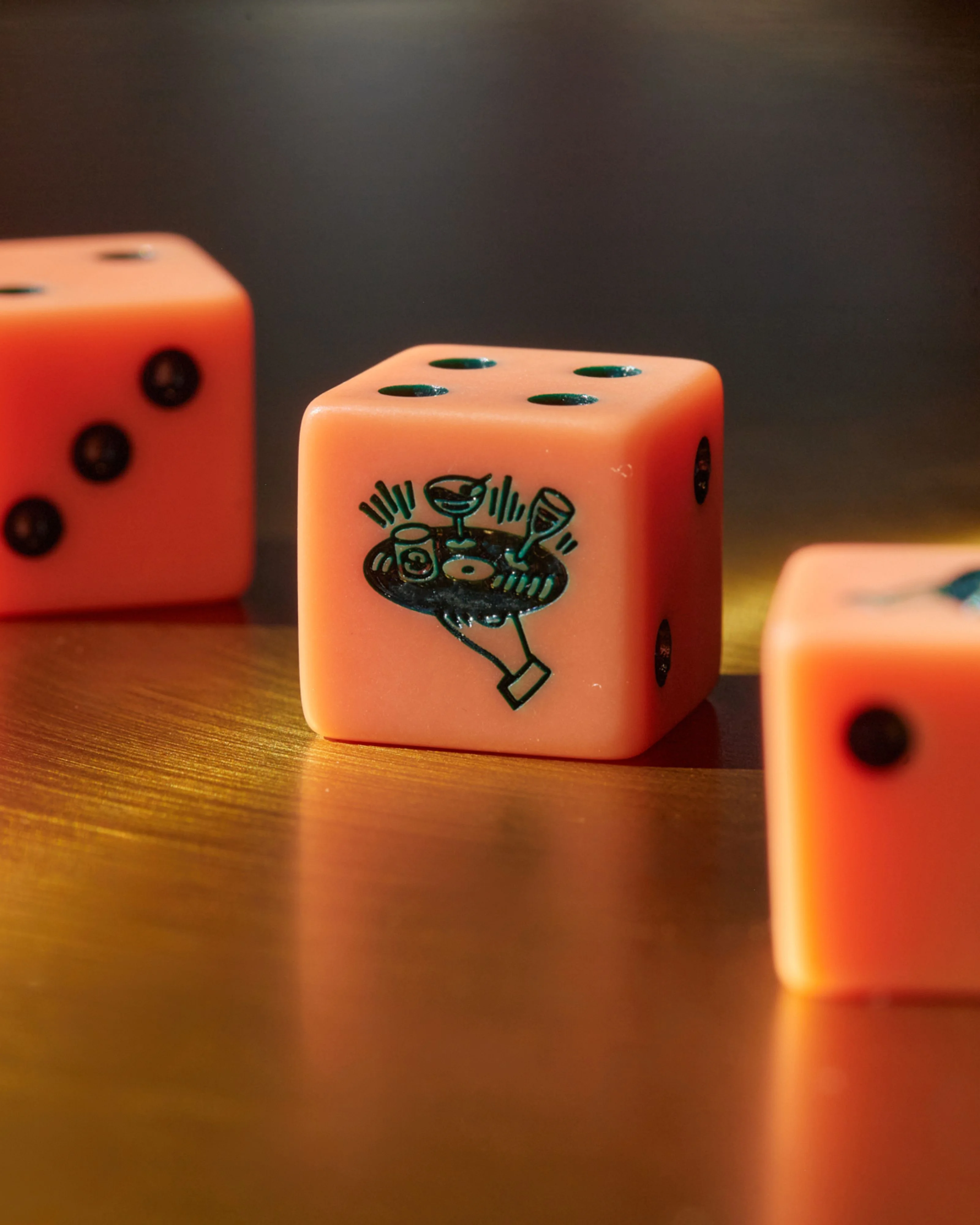 Bright orange dice arranged on a warm wooden surface, with the center die featuring a custom illustration of a decorative vessel or urn with black line details, while surrounding dice display traditional pip markings in dark teal.