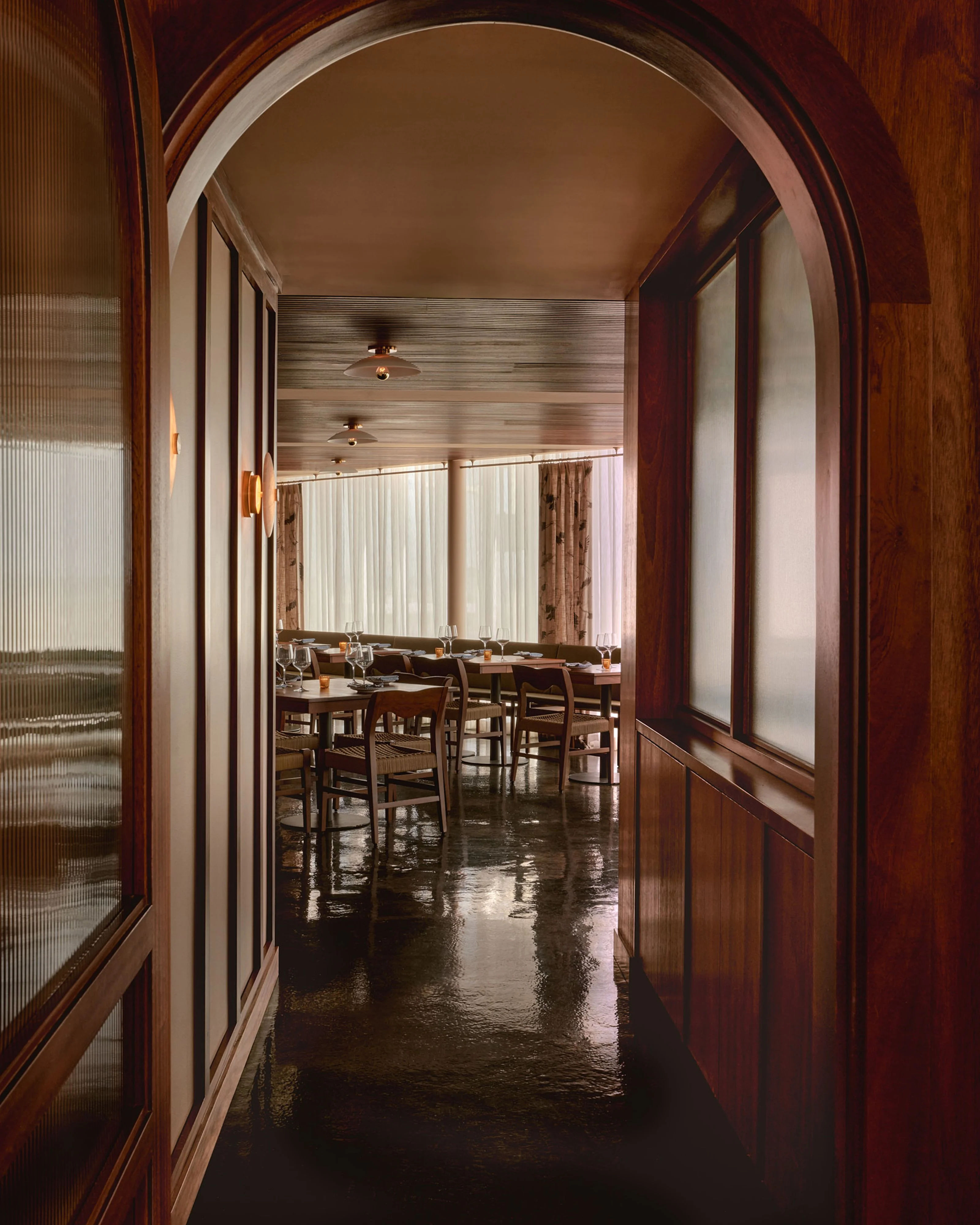 A warm, modern dining space framed by a striking arched doorway with rich burgundy wooden doors and frosted glass panels; the view extends through to a sleek dining room featuring dark polished concrete flooring, wooden chairs, and floor-to-ceiling vertical blinds that filter soft natural light.