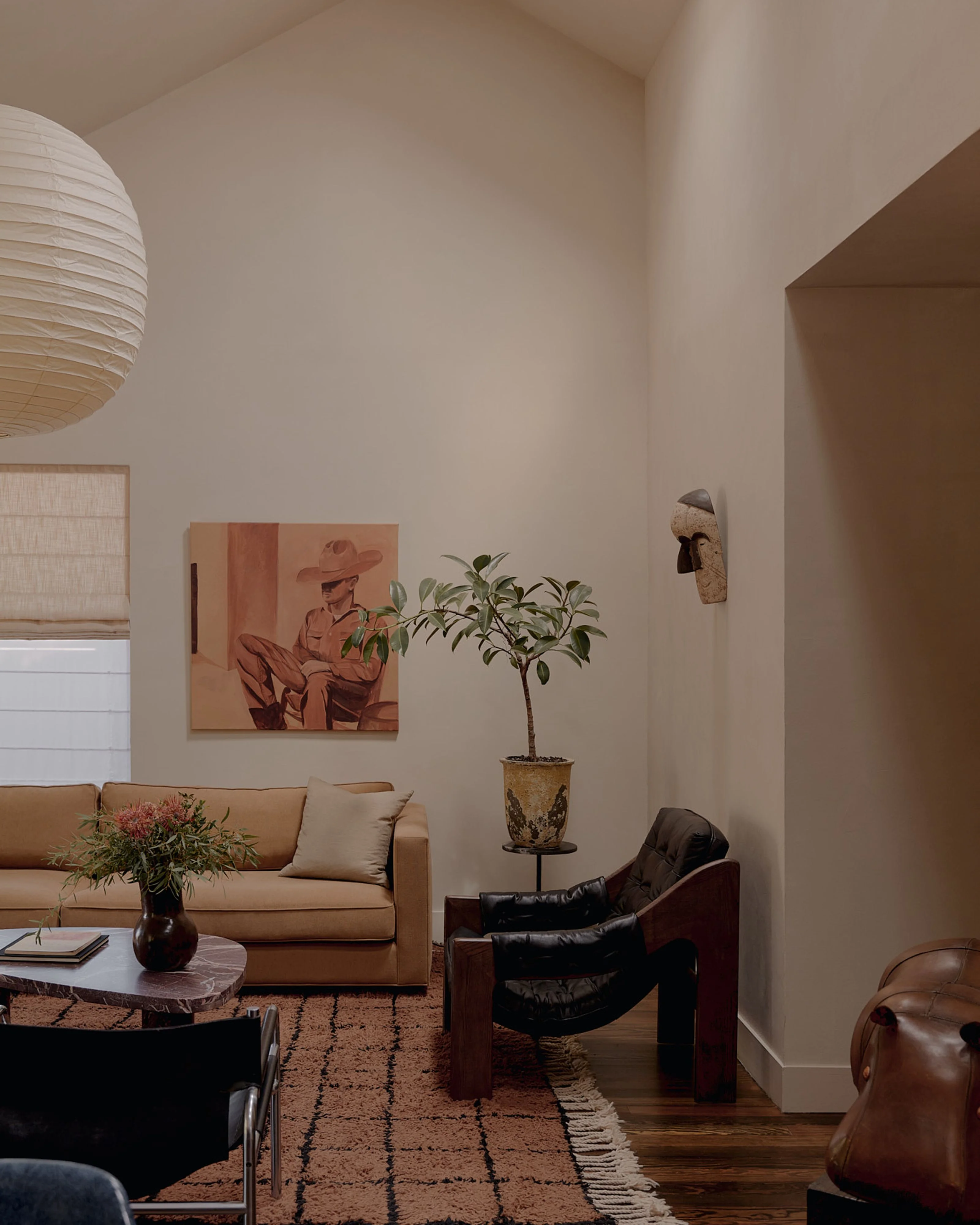 A minimalist living room bathed in natural light from a skylight features cream-colored walls, warm wood flooring, and a curated collection of mid-century modern furniture including a tan linen sofa, black leather lounge chair, and wooden side table with potted plant.