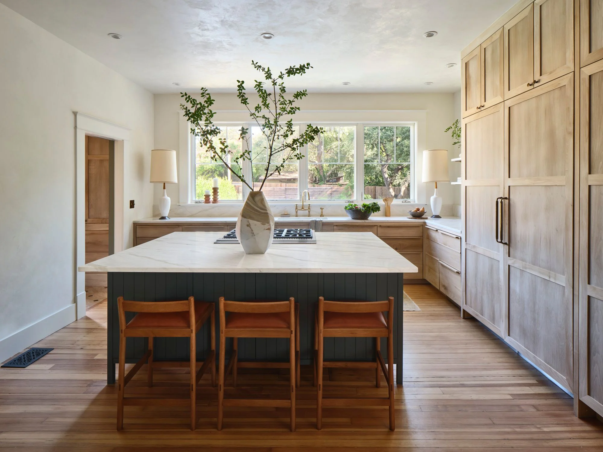A bright, modern kitchen features a central island with a black vertical-paneled base and white marble countertop, surrounded by three mid-century wooden chairs, while light wood cabinetry lines the perimeter and large windows flood the space with natural light overlooking a garden.