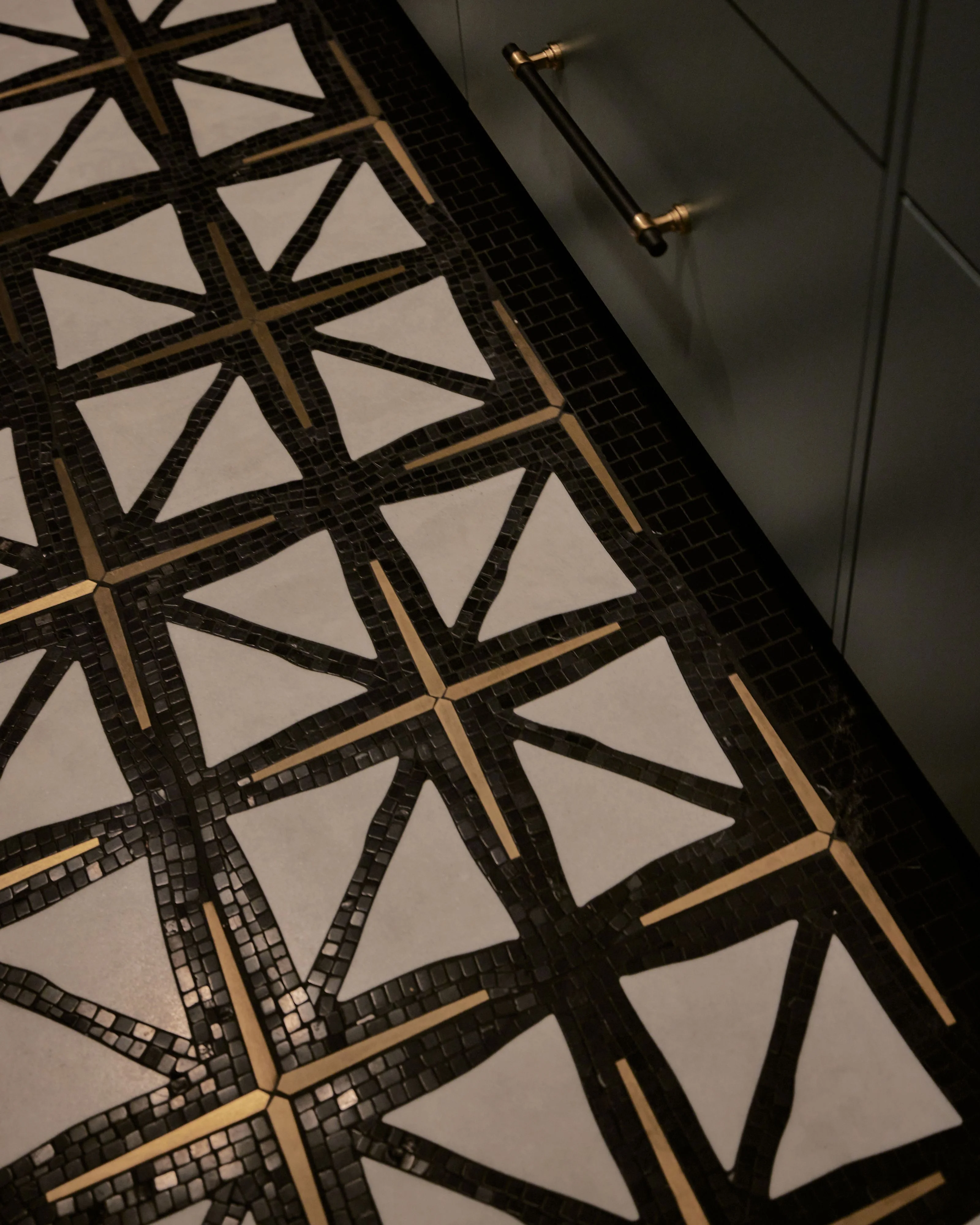 A modern interior features a striking geometric ceiling design with black mosaic tiles and brass accents forming angular patterns against a pale cream background, complemented by a sleek gray wall with brass hardware below.