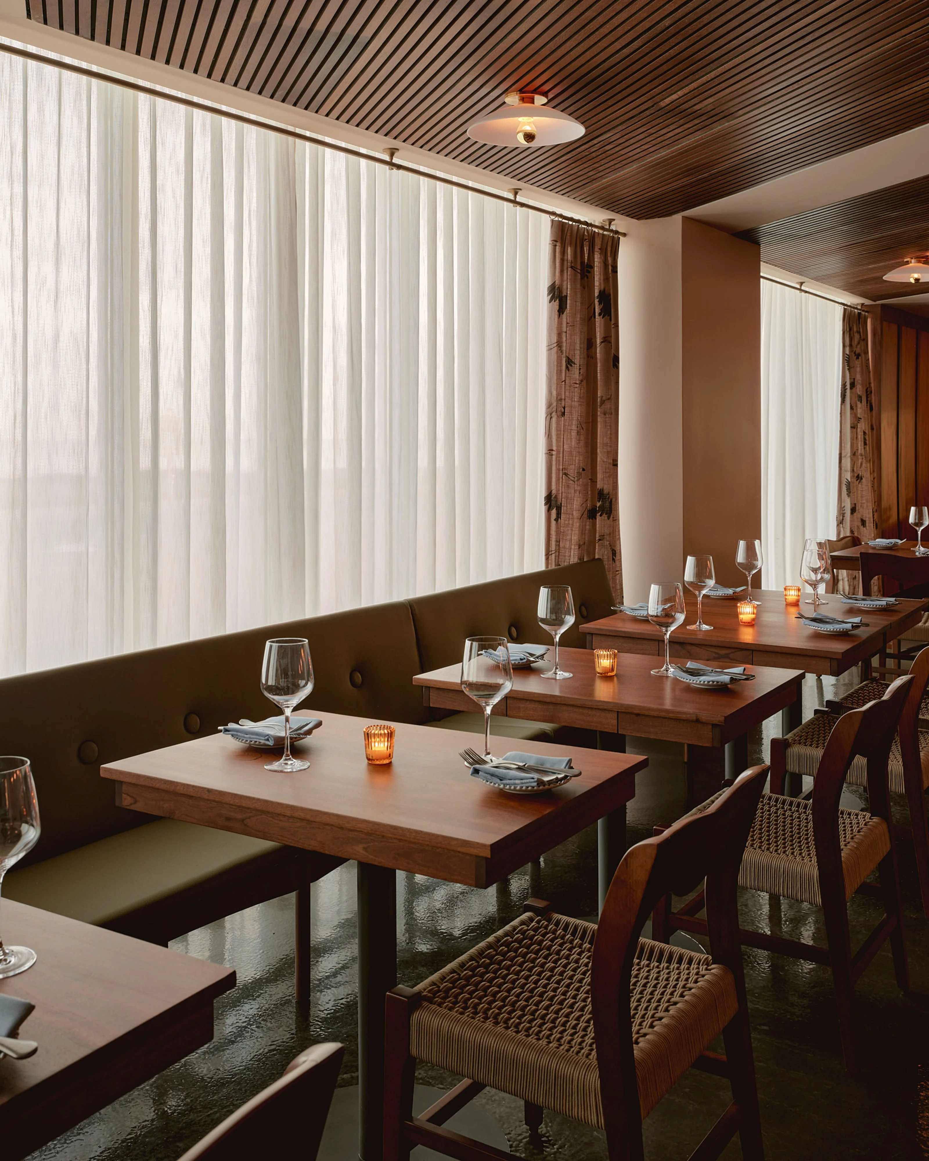 A modern fine dining restaurant features wooden tables with olive-green banquette seating arranged along floor-to-ceiling vertical blinds that flood the space with diffused natural light. The sophisticated interior incorporates a striking slatted wood ceiling, warm brown tones, woven chair details, amber candlelight, and place settings with wine glasses.