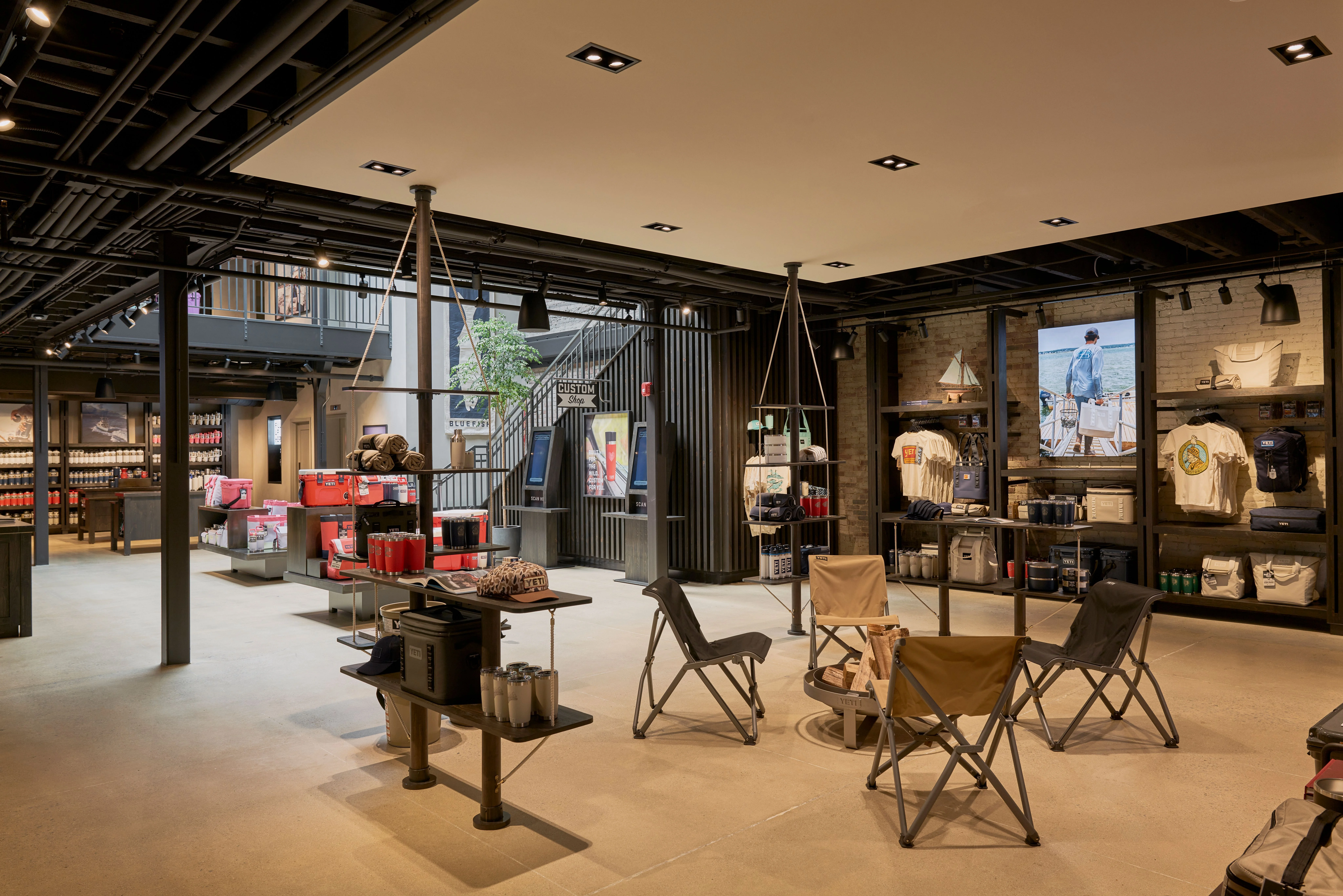 A modern retail showroom featuring an industrial-meets-outdoor aesthetic, with exposed steel beams, concrete flooring, and a neutral color palette of tans and blacks, displaying camping gear and apparel across shelving units and display tables arranged with camping chairs and a central fire pit installation.