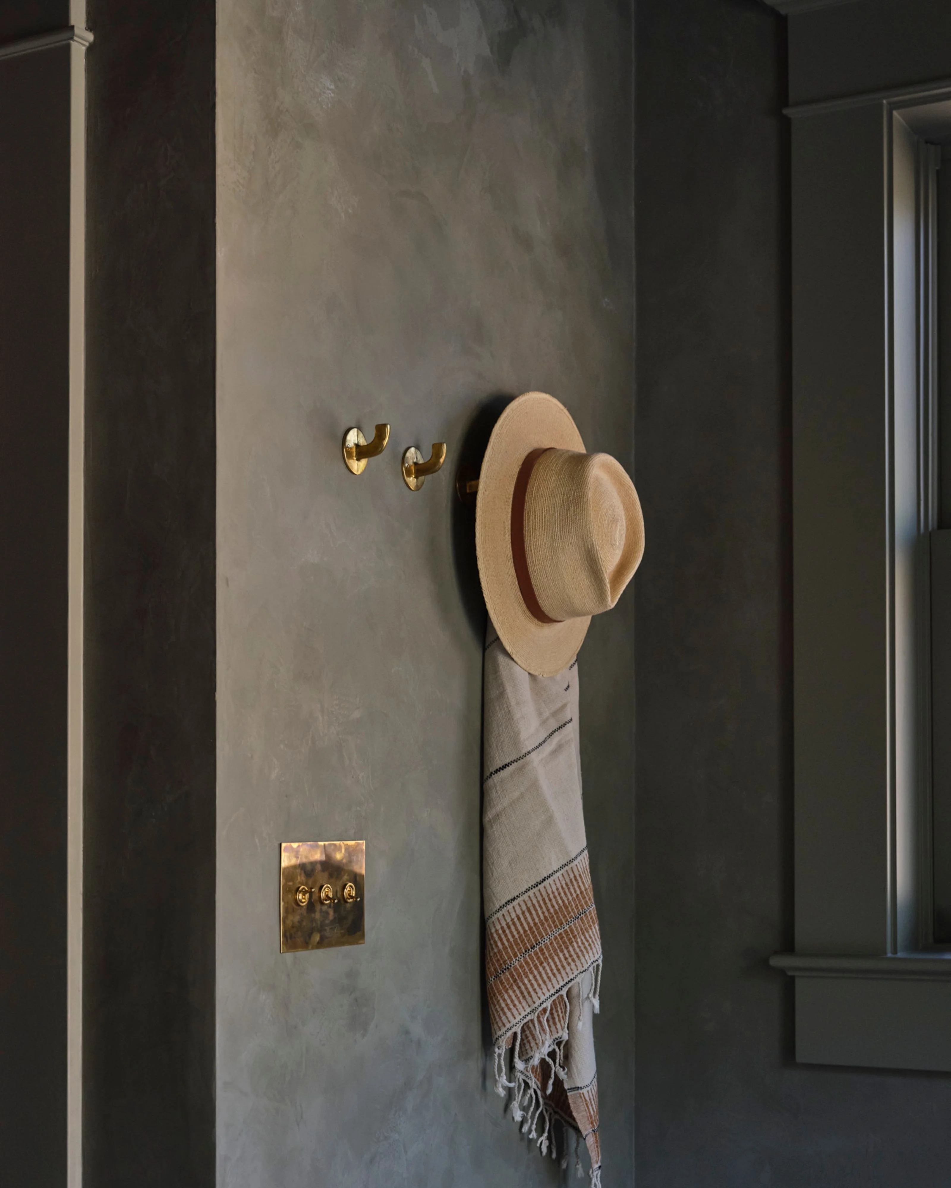 A minimalist entryway features textured concrete walls in muted gray tones, accented with polished brass fixtures including small wall hooks and an ornate switch plate; a natural woven straw hat and striped linen cloth hang from a brass hook, creating a serene, contemporary aesthetic.
