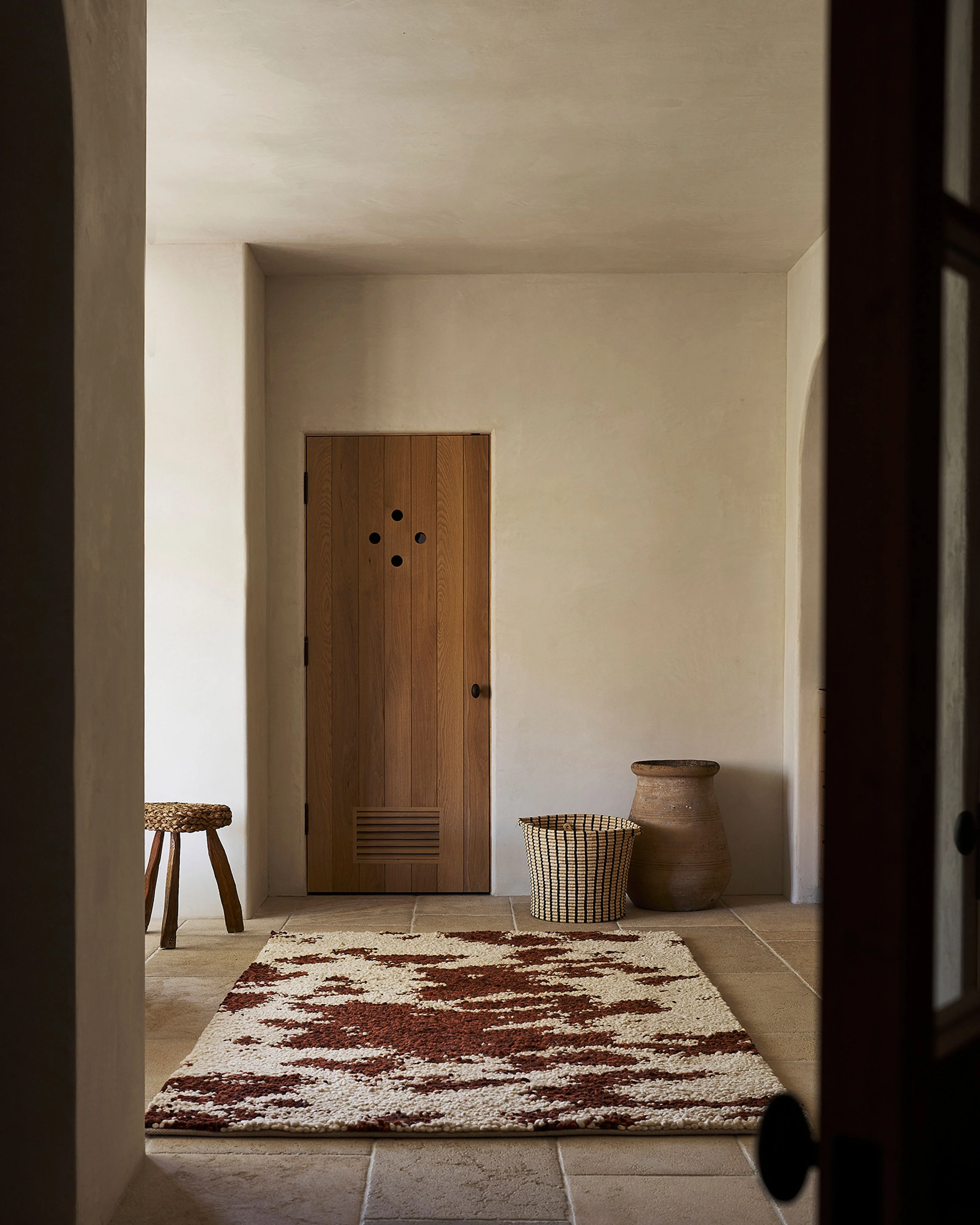 A minimalist interior hallway with cream-colored plaster walls and stone flooring, featuring a wooden door with circular hardware, a woven stool, textured baskets, and a terracotta vessel arranged sparsely against the walls, unified by a cream and burgundy patterned rug.