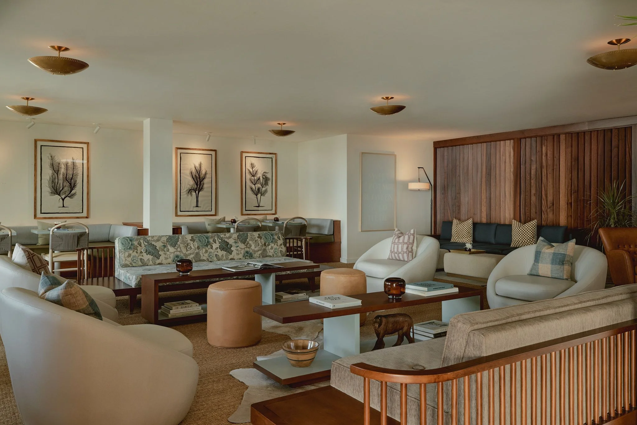 A mid-century modern hotel lounge featuring curved cream-colored seating, warm wood furniture, and brass disc ceiling lights arranged throughout the spacious room. The neutral palette is accented by botanical artwork in wood frames, patterned upholstery, and rich wood paneling on the right wall.