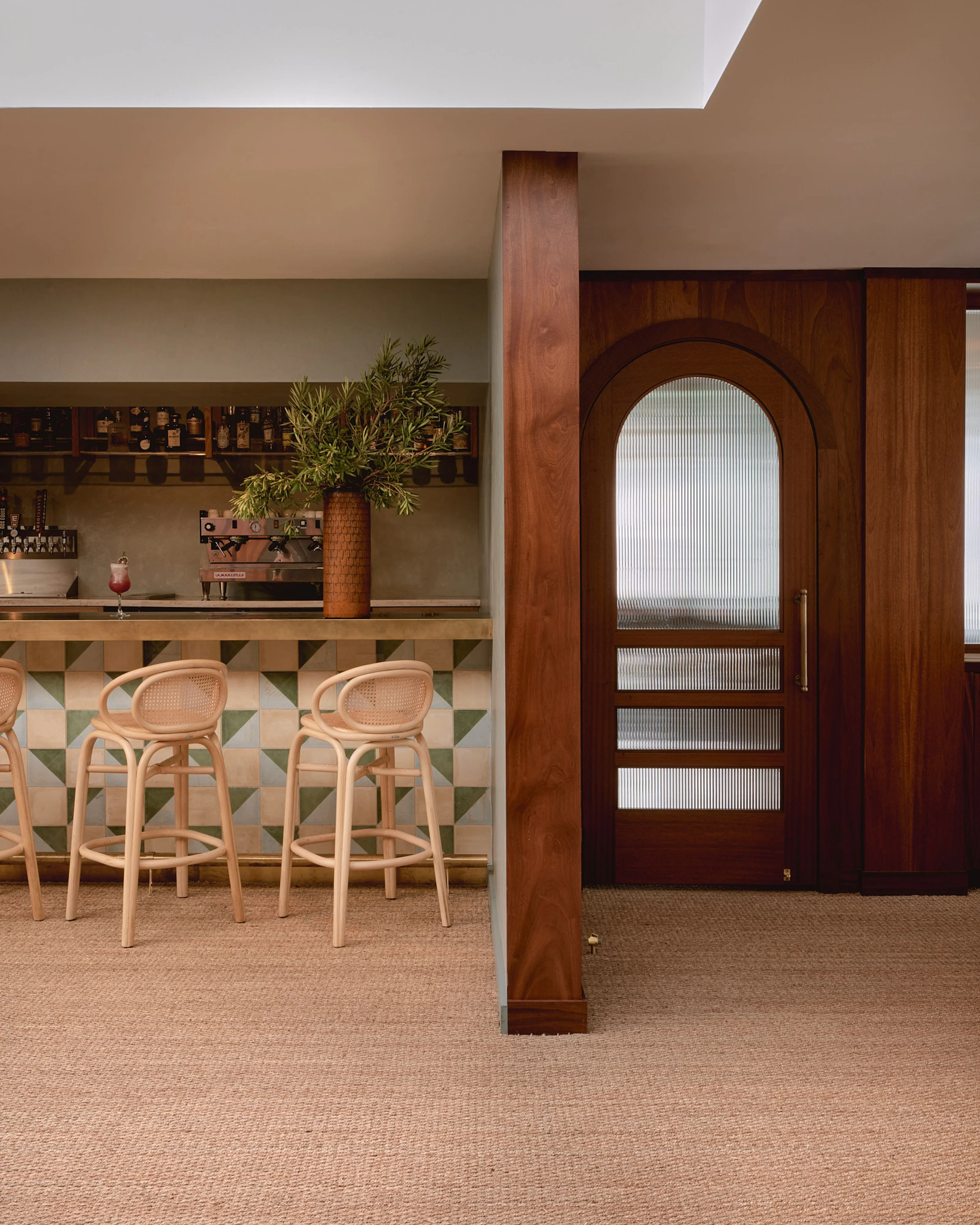 A mid-century modern bar and lounge space features warm taupe walls, natural light wood furnishings, and woven rattan bar stools arranged along a geometric tile-patterned bar counter, while a striking dark wood arched doorway with frosted glass panels serves as a focal point, flanked by a prominent wooden column.