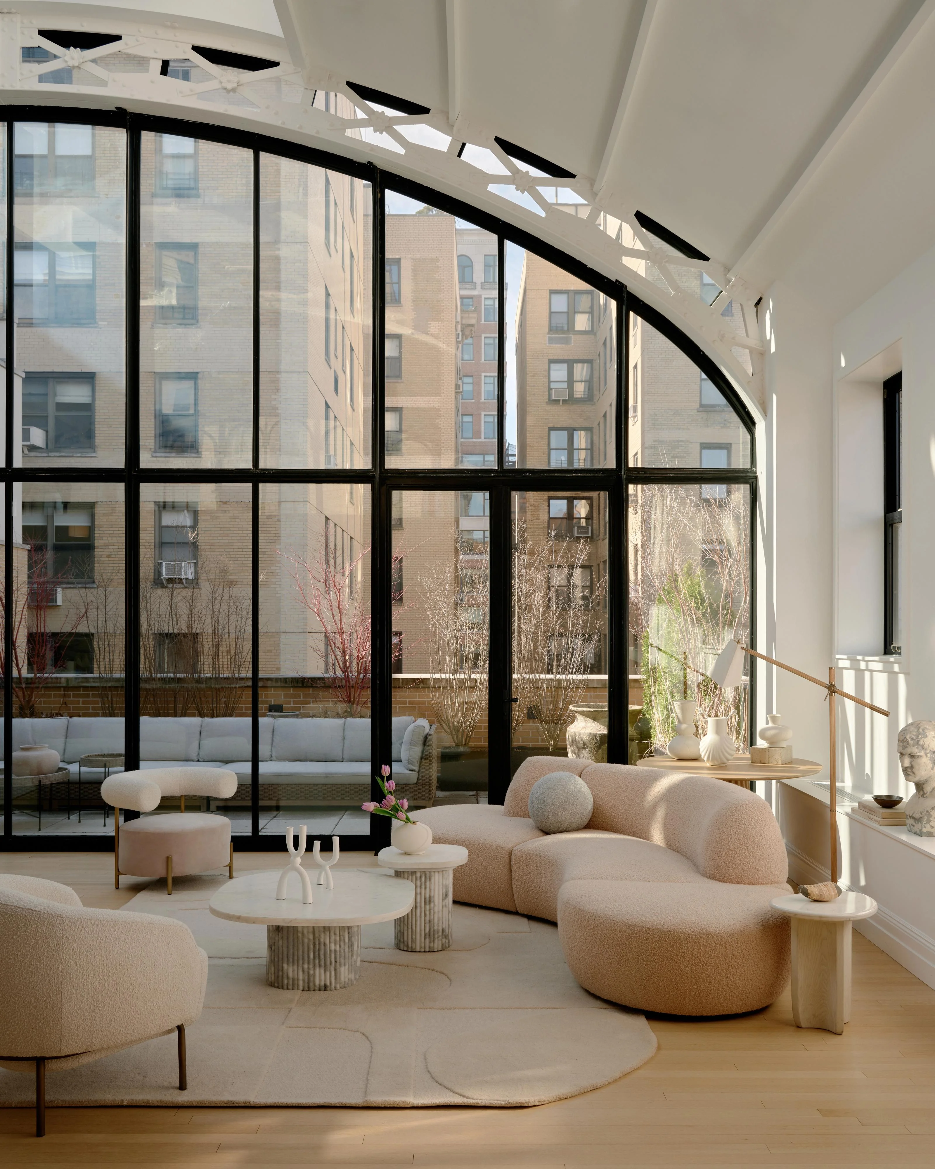 A bright, minimalist living room features curved, cream-colored upholstered furniture arranged on light wood flooring, accented by cylindrical side tables and a geometric area rug. Floor-to-ceiling black-framed windows with a curved top and skylights flood the space with natural light, overlooking an urban courtyard.